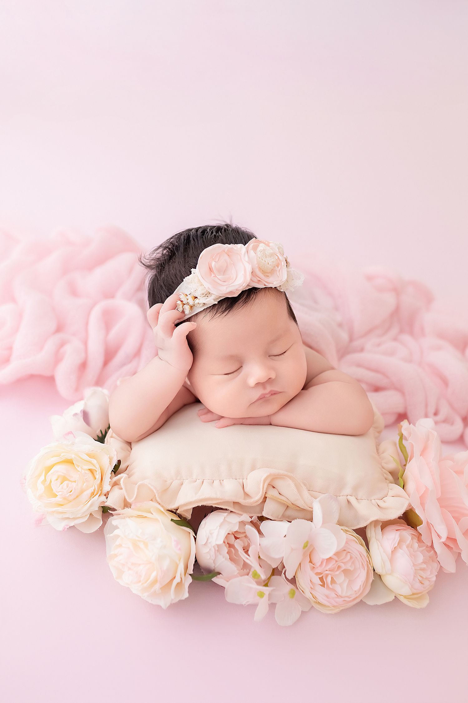 Sleeping newborn baby girl with floral headband resting on cream pillow surrounded by pink roses.