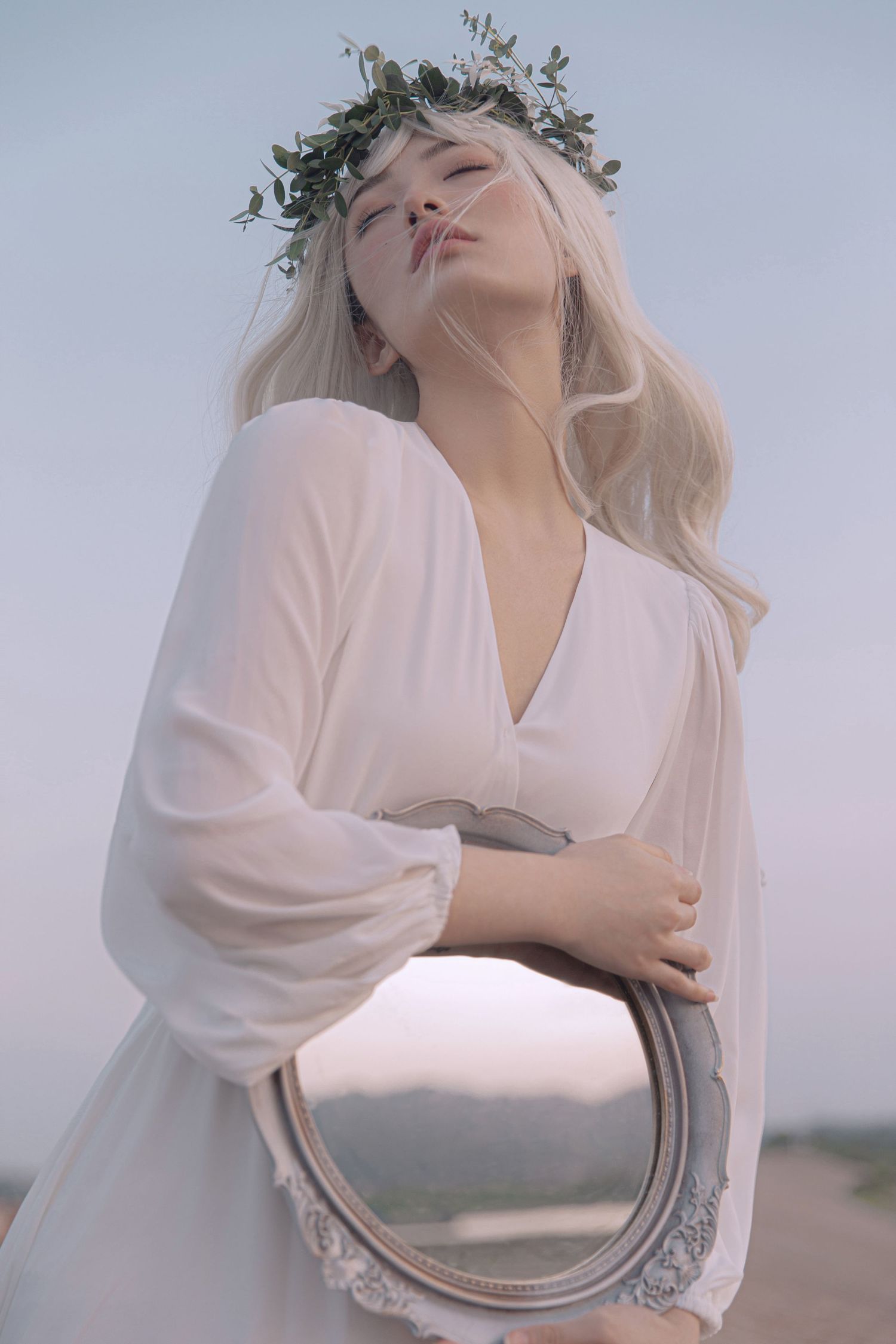 Blonde woman in white dress holding oval mirror with eyes closed, wearing green leaf crown outdoors.