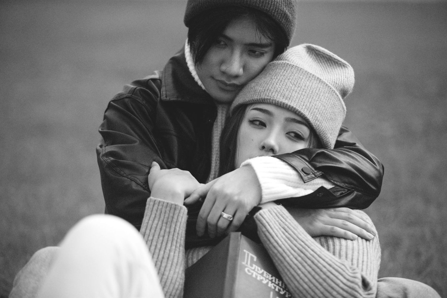 Black and white photo of a young Asian couple sitting on grass, embracing tenderly in cozy winter knitwear.