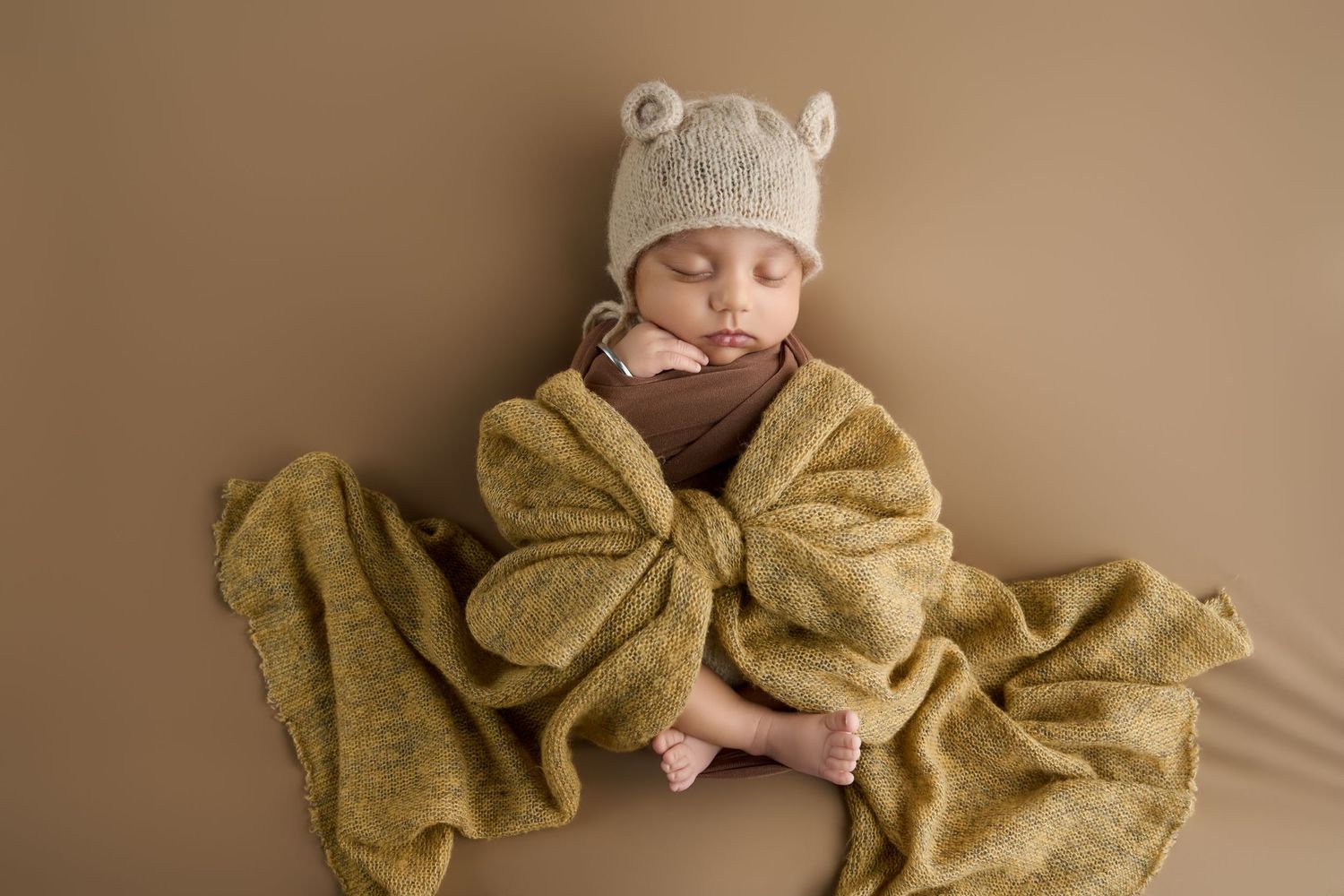 Newborn baby sleeping peacefully wrapped in a golden knit blanket wearing a bear ear hat on a tan background.