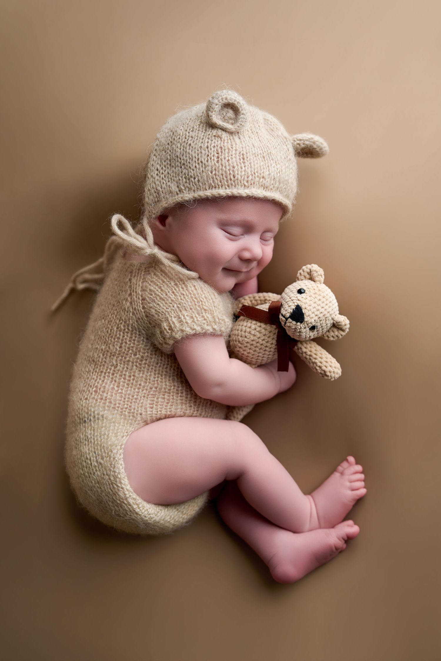 Sleeping newborn baby in beige knit bear outfit hugging a small crocheted teddy bear on tan background.