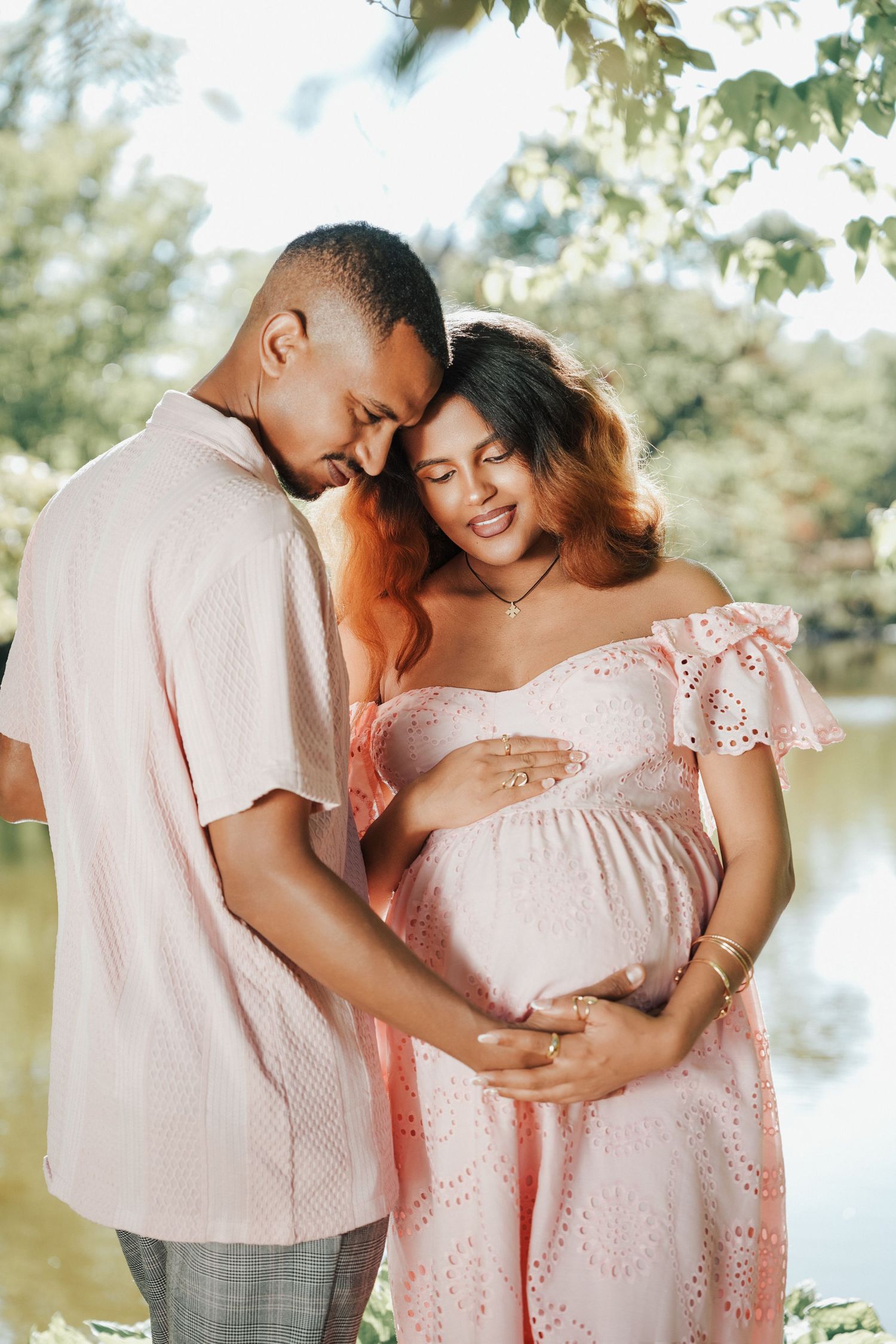 A couple tenderly cradles a pregnant belly outdoors near a pond, both wearing matching pink outfits in a maternity photo.