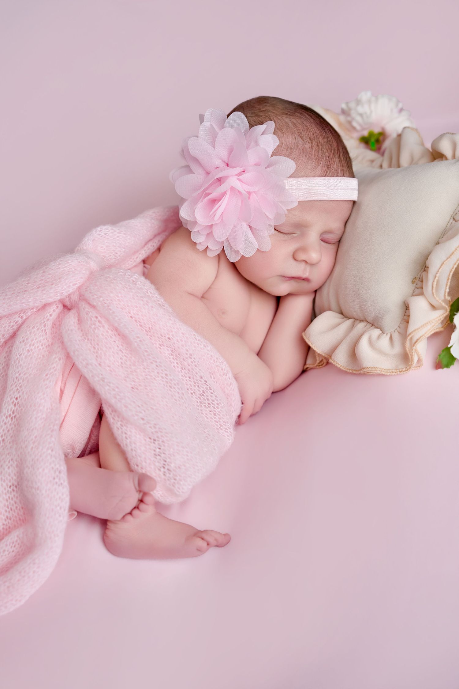 Sleeping newborn baby girl wearing a pink flower headband, wrapped in a soft pink knit blanket on a pink background.