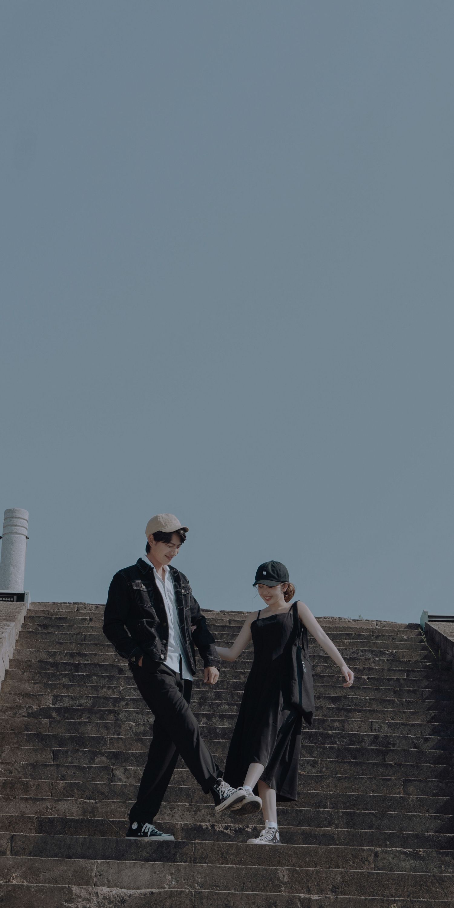 A young couple in black outfits descending outdoor stone steps under a clear blue sky.