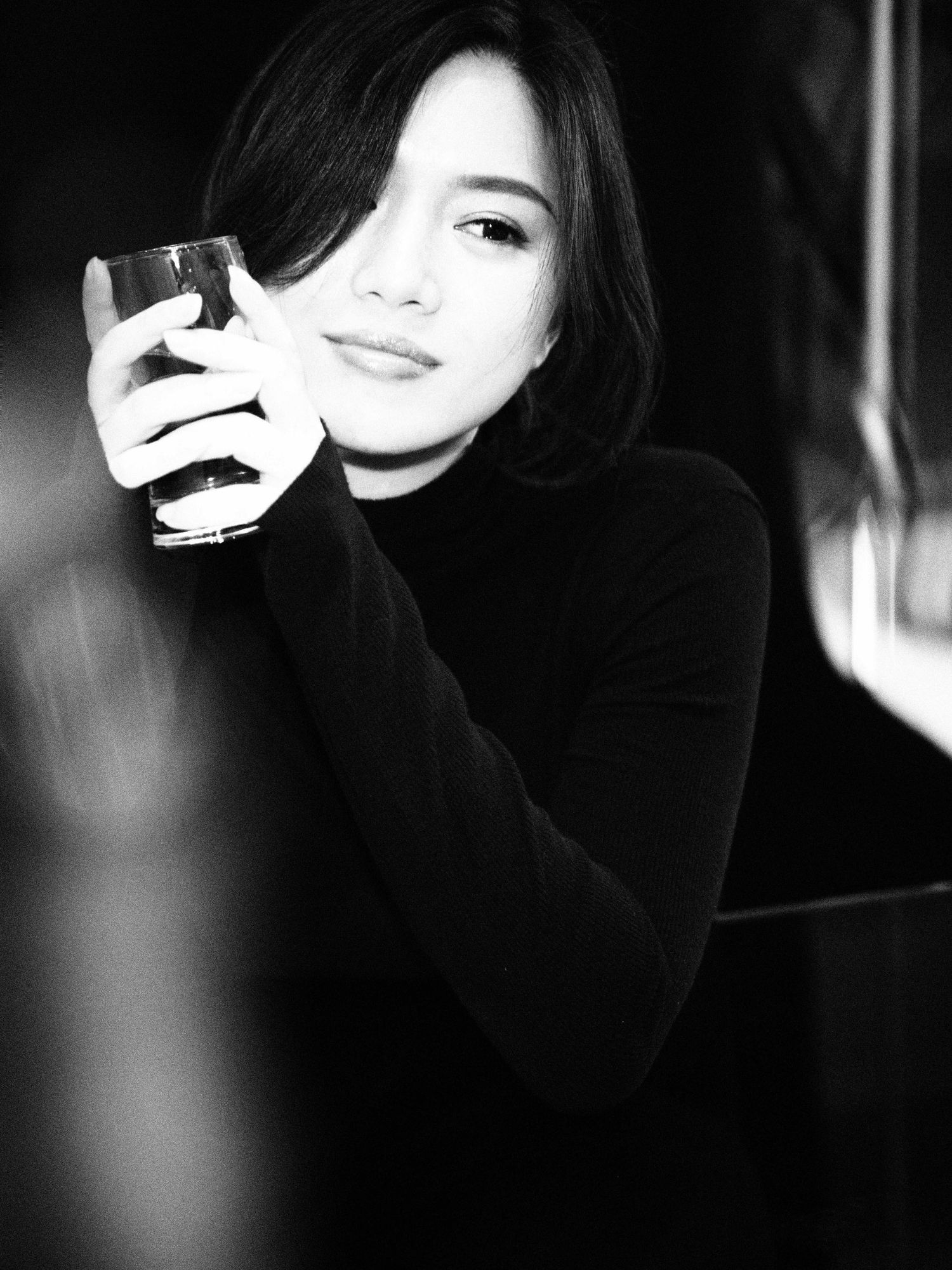 Black and white portrait of an Asian woman in a turtleneck holding a glass, with a soft smile.
