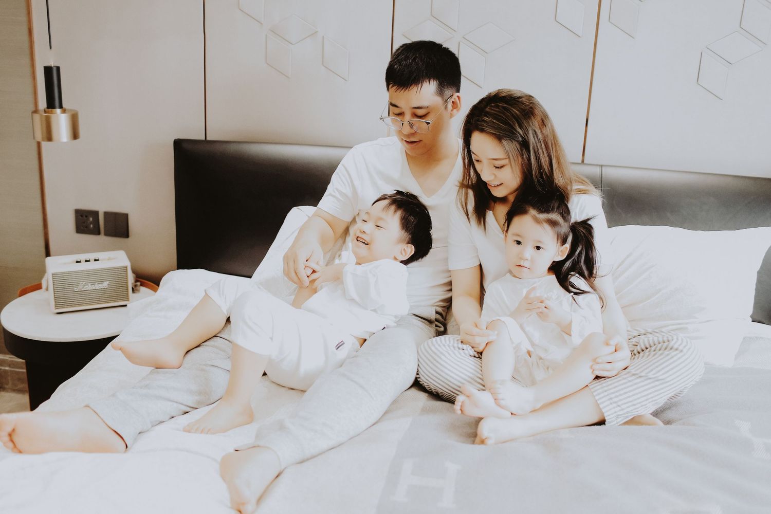 An Asian family of four sits together on a white bed, parents and two young children sharing a calm moment.