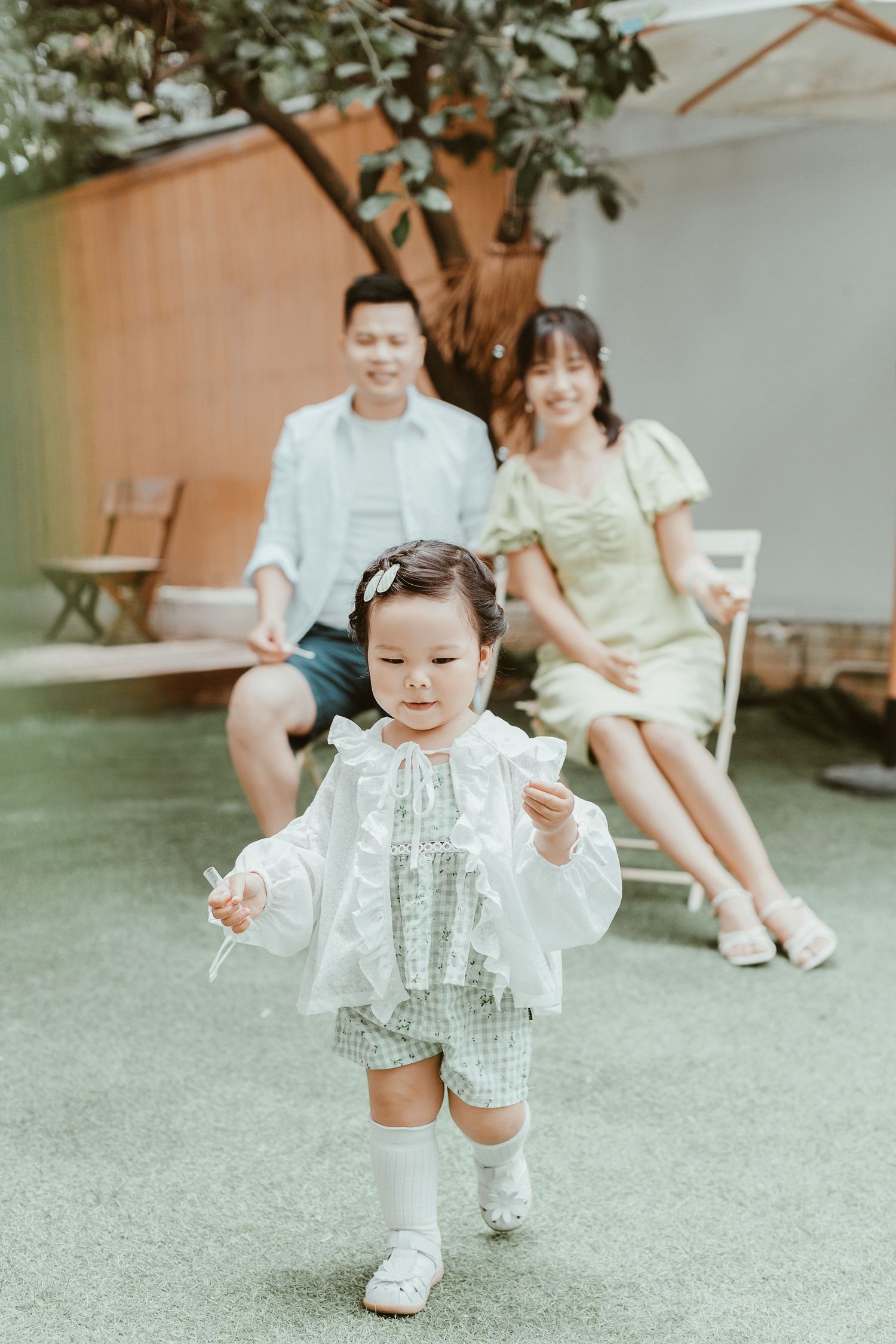 A young Asian toddler girl in a white ruffled outfit walks outdoors while her smiling parents sit together in the background.