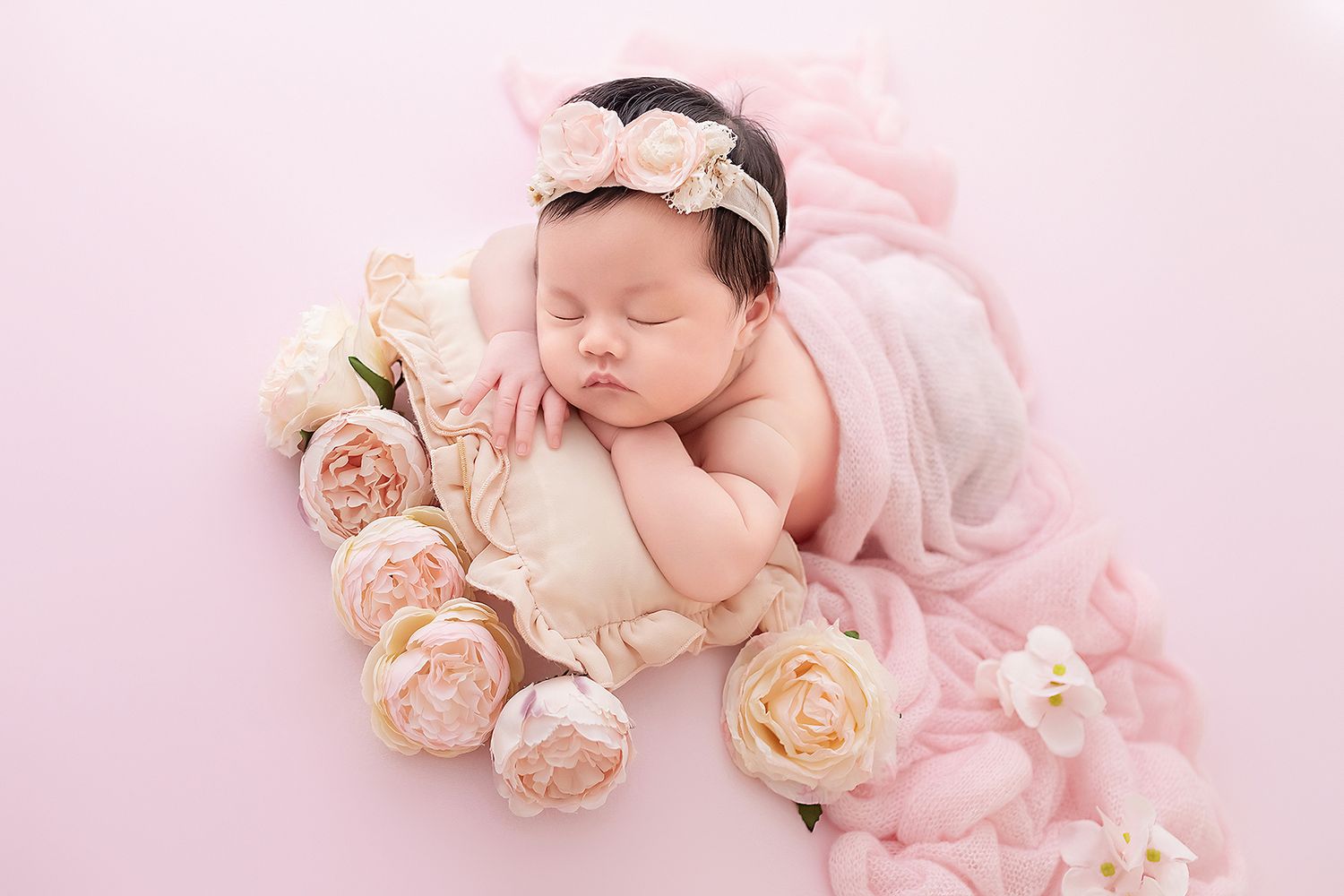 Newborn baby girl sleeping on pillow wrapped in pink blanket surrounded by peony flowers.