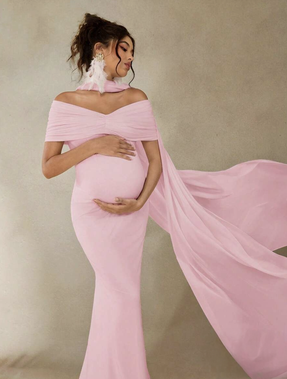 Pregnant woman in elegant pink off-shoulder mermaid maternity gown with flowing chiffon cape for photoshoot.