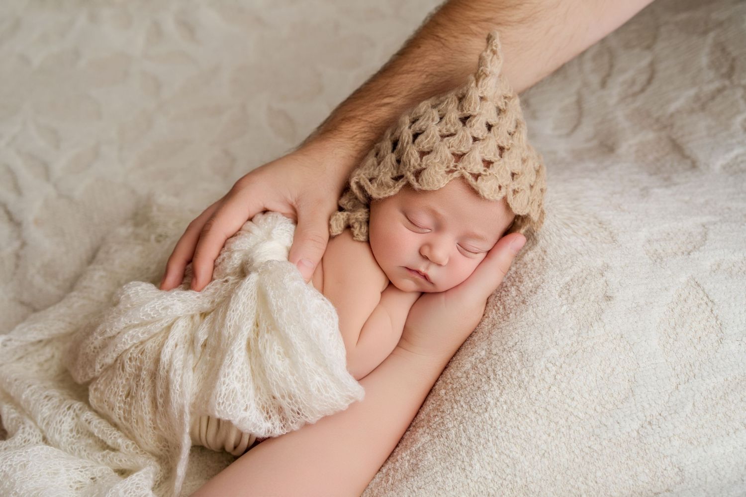 Sleeping newborn baby wearing a knit beige hat, cradled gently by an adult hand on a soft cream blanket.