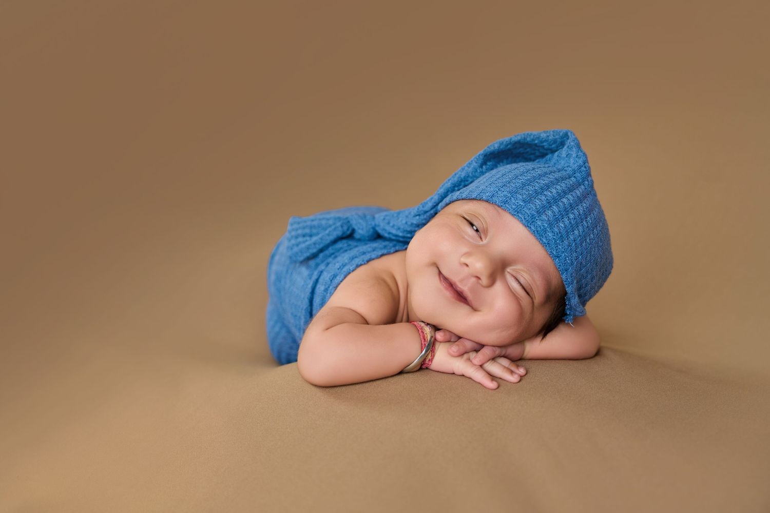 Smiling newborn baby wearing a blue knit hat and wrap, lying on a tan background in a professional photo.