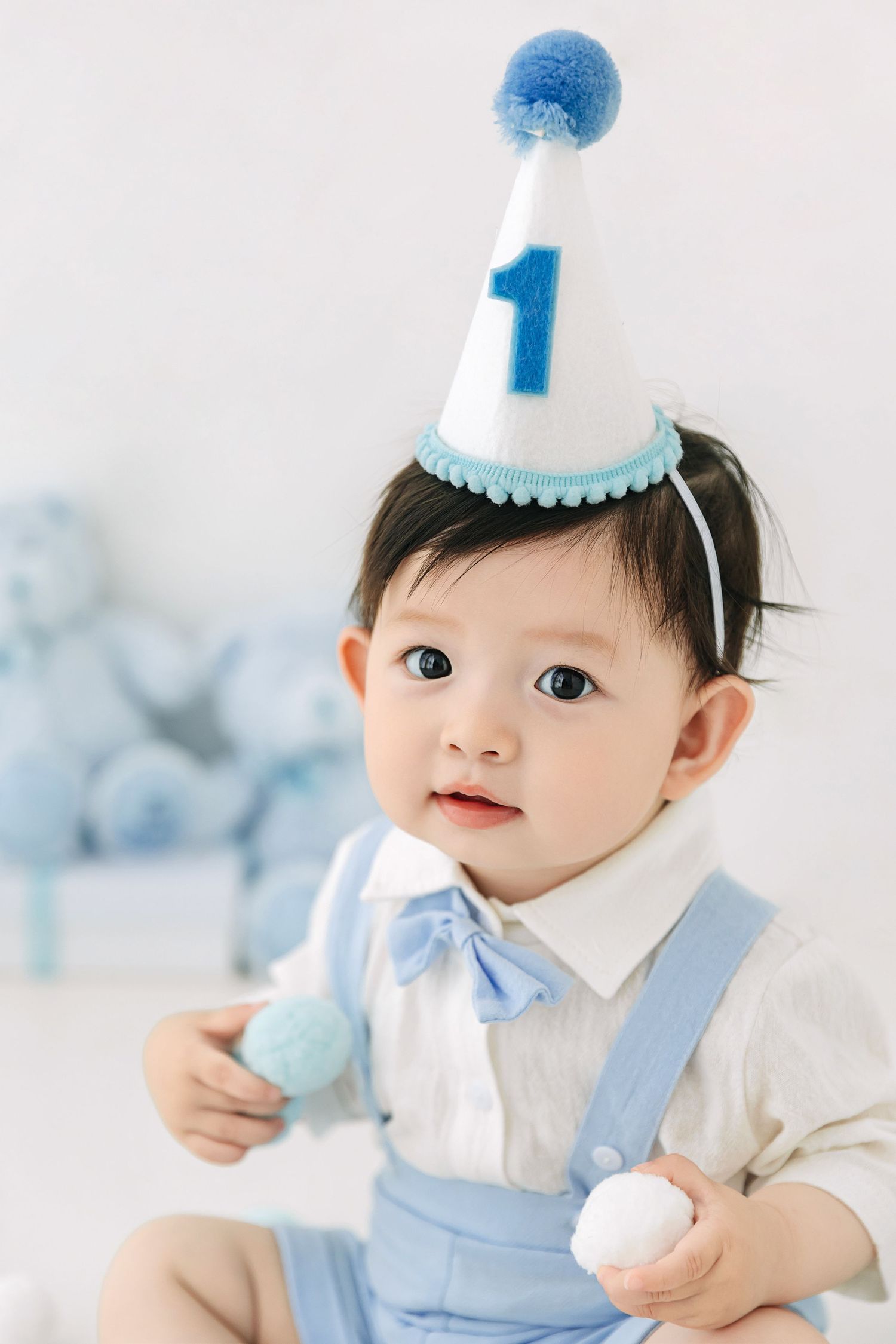 A baby boy wearing a blue number 1 birthday hat and suspenders holds treats during his first birthday photo shoot.