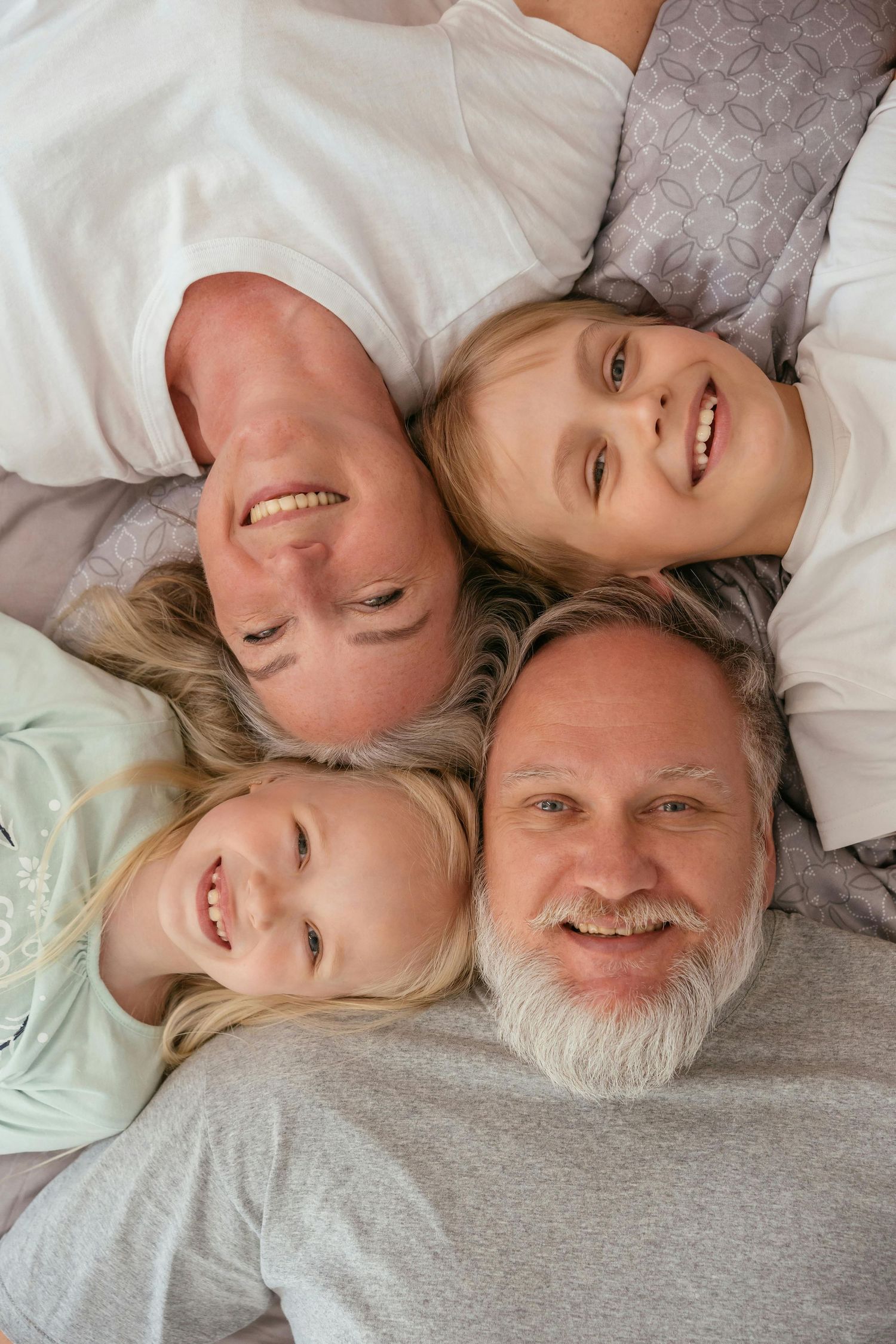 Happy family of four lying together on a bed, smiling at camera from above in cozy pajamas.