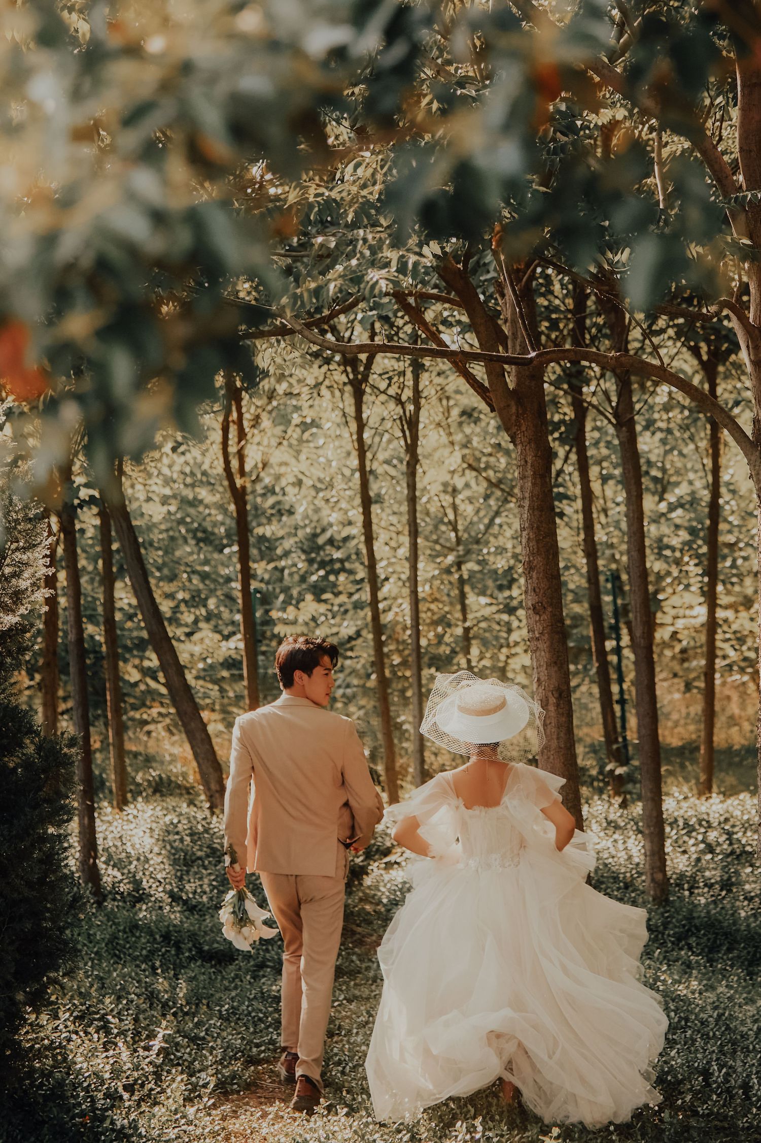 Newlywed Asian couple walks hand-in-hand through a sunlit forest, bride in white ballgown and wide-brim hat.