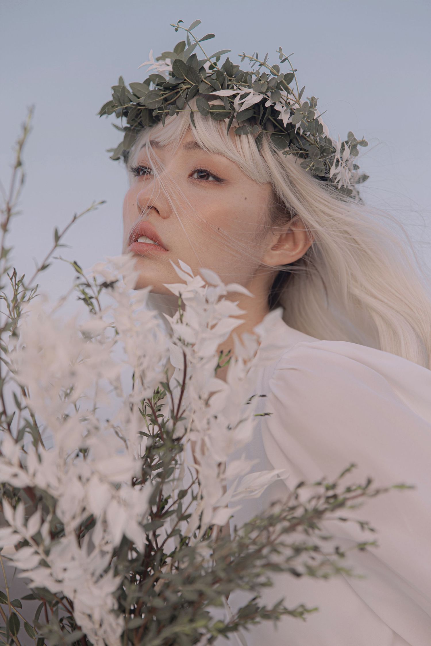 Ethereal bride with floral crown and blonde hair holding white wildflowers against a soft blue sky.