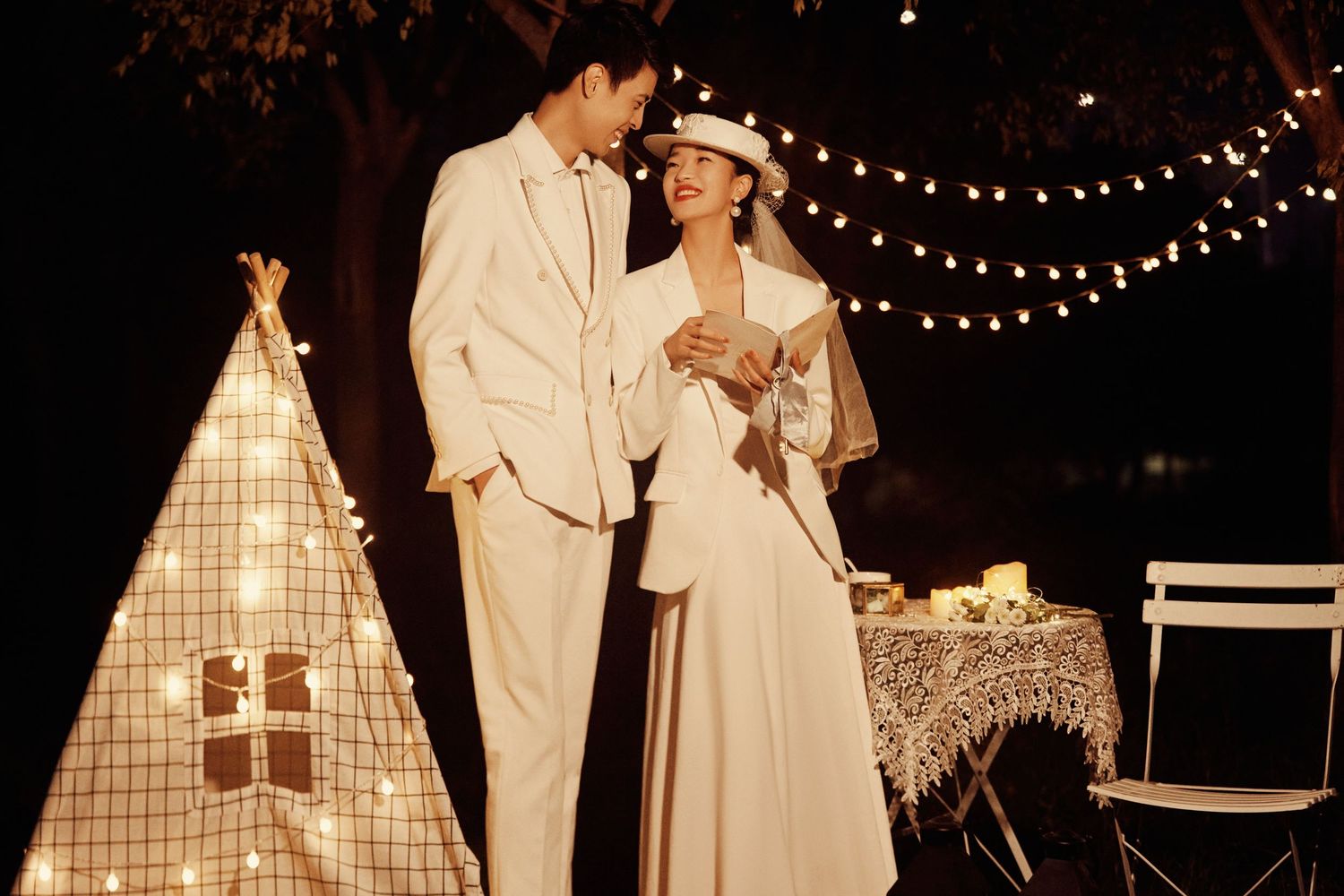 Romantic couple in elegant white wedding attire stand together near glowing teepee with string lights at night.