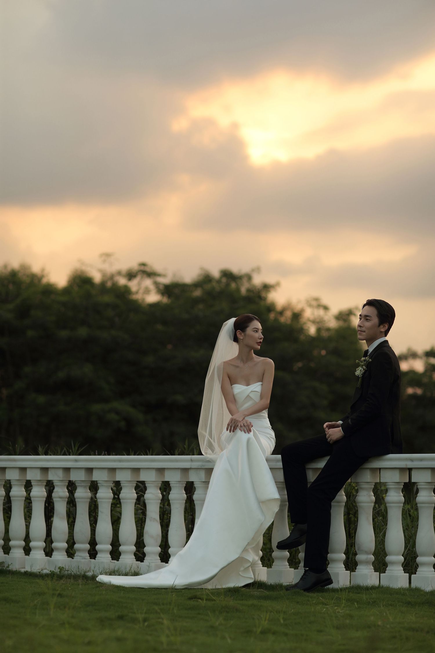 Bride and groom sit on white stone balcony railing smiling together during dramatic sunset sky.