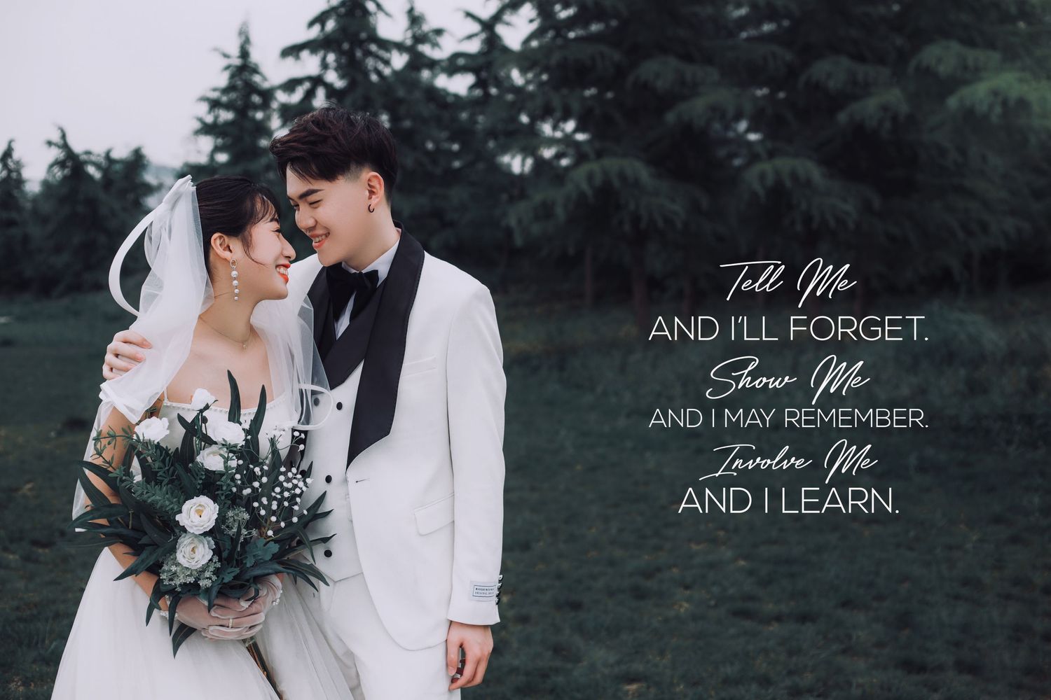 Romantic couple in wedding attire share intimate moment holding bouquet with evergreen background.