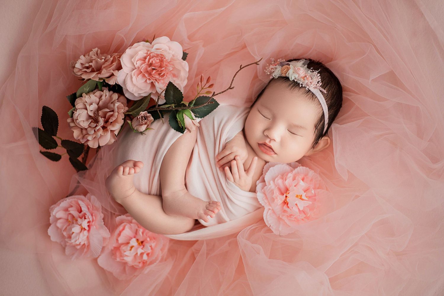 Sleeping newborn baby girl in pink wrap with floral headband nestled in bowl surrounded by pink peonies.