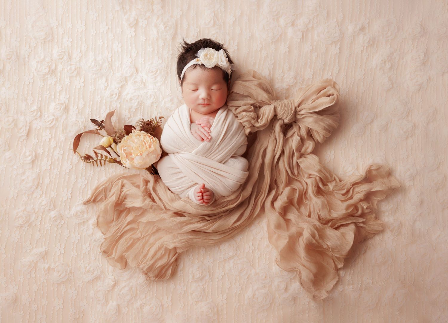 Sleeping swaddled newborn baby girl with floral headband on cream textured blanket.