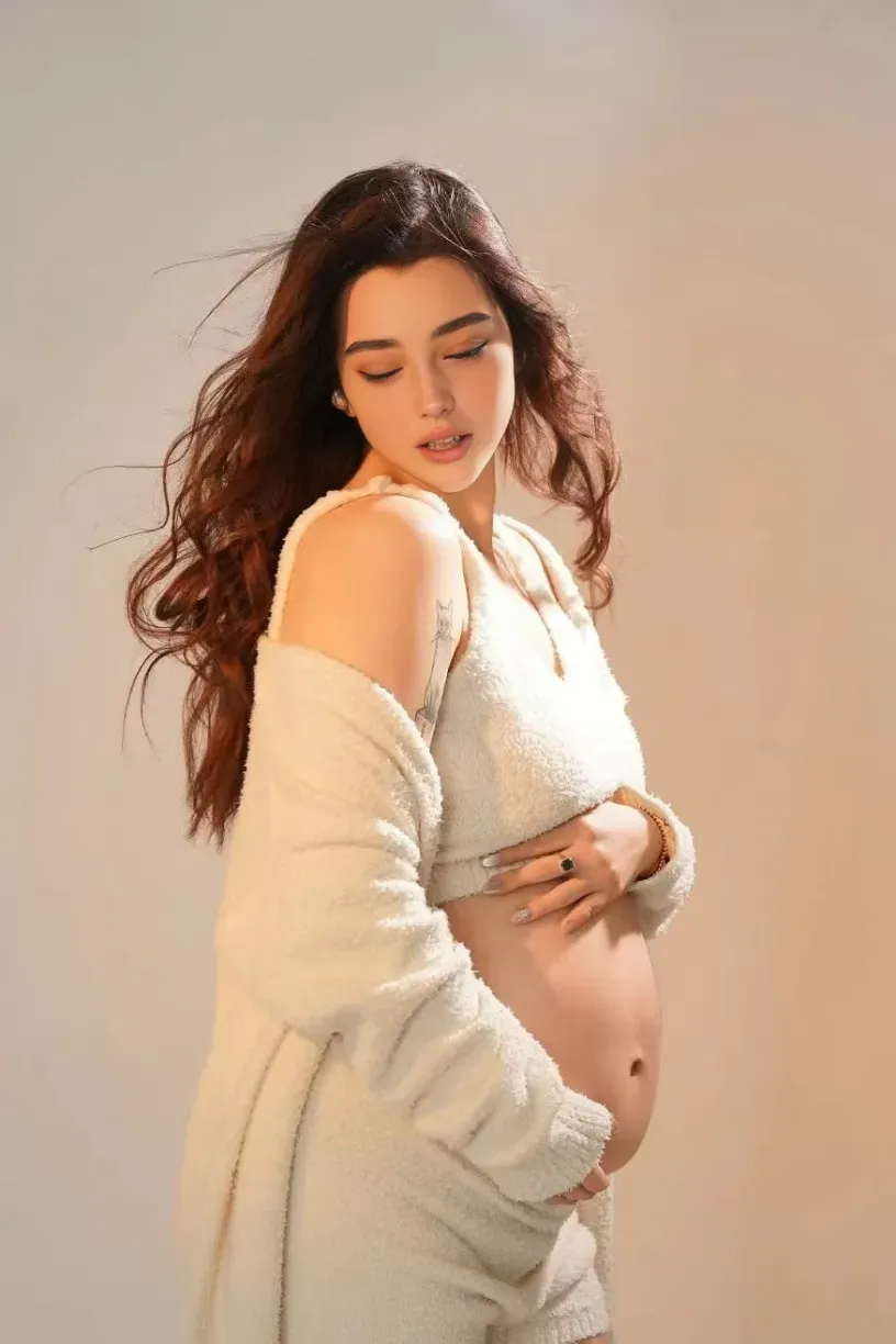 Pregnant woman in white fluffy cardigan and crop top cradling baby bump in warm-toned studio setting.