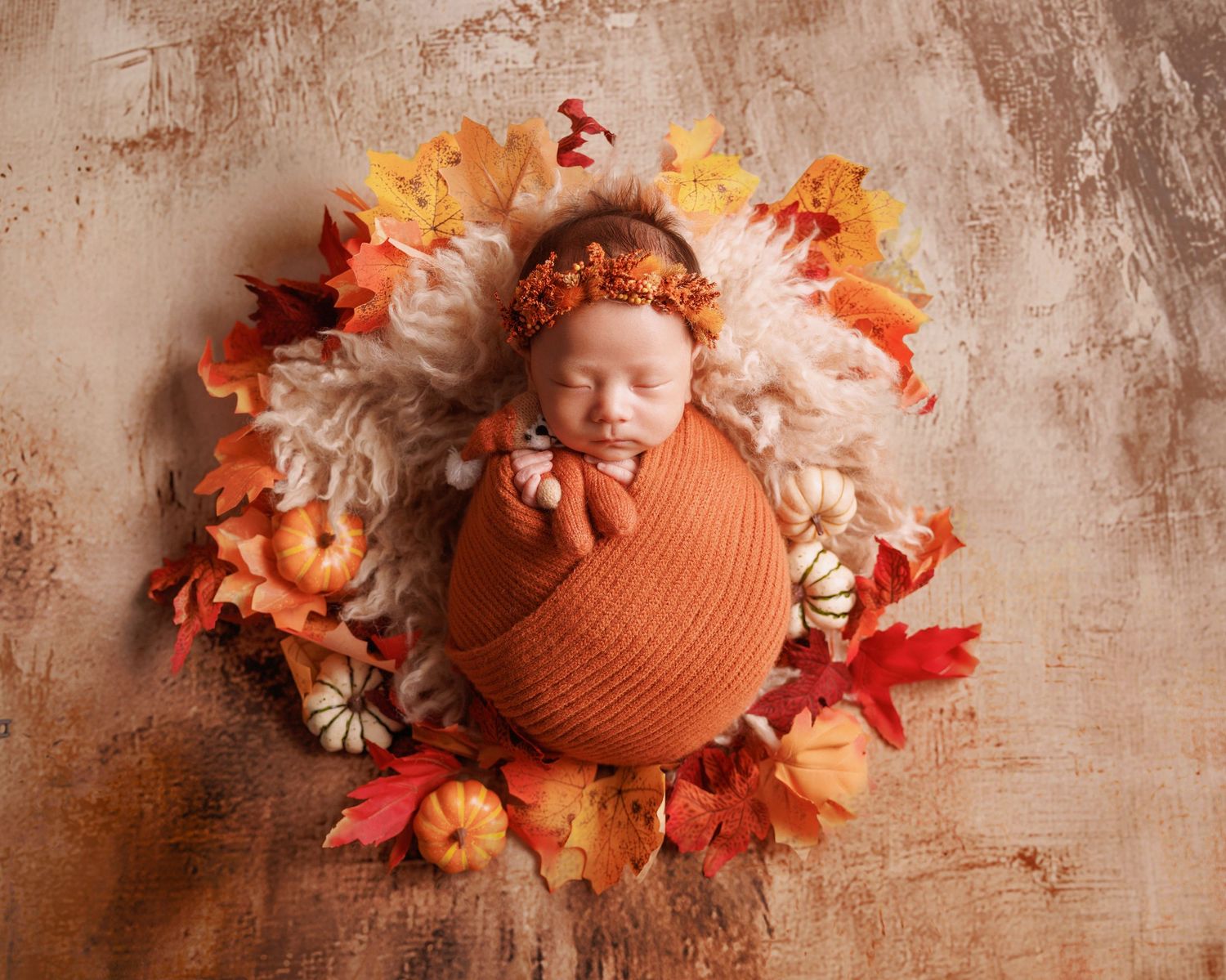 Swaddled newborn wearing orange floral headband, peacefully sleeping in a fall-themed arrangement with pumpkins.