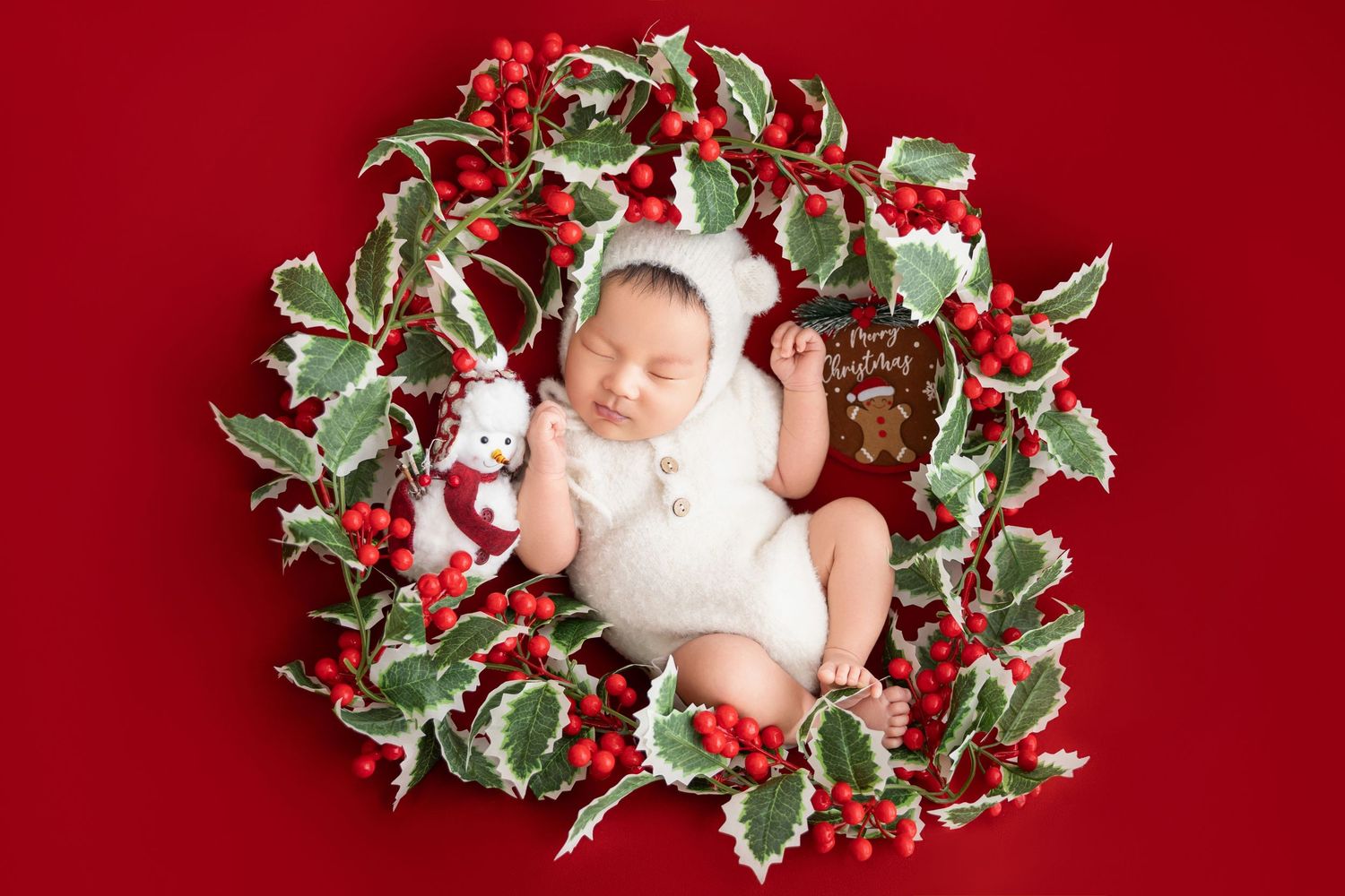 Newborn baby in white bear bonnet sleeping inside circular holly wreath with red berries on festive red background.