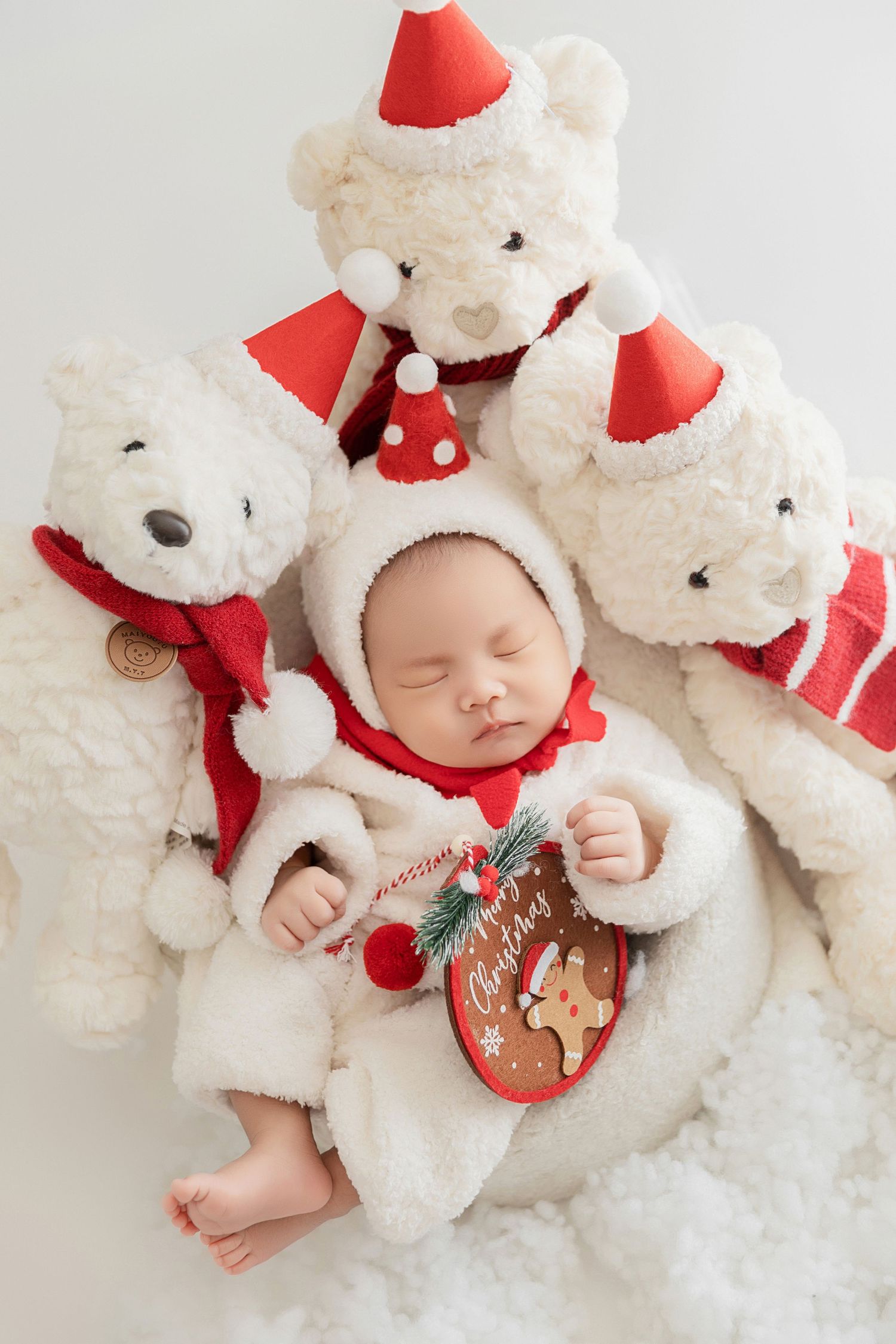 Sleeping newborn in white fluffy Santa outfit surrounded by white teddy bears in Christmas hats on snowy backdrop.