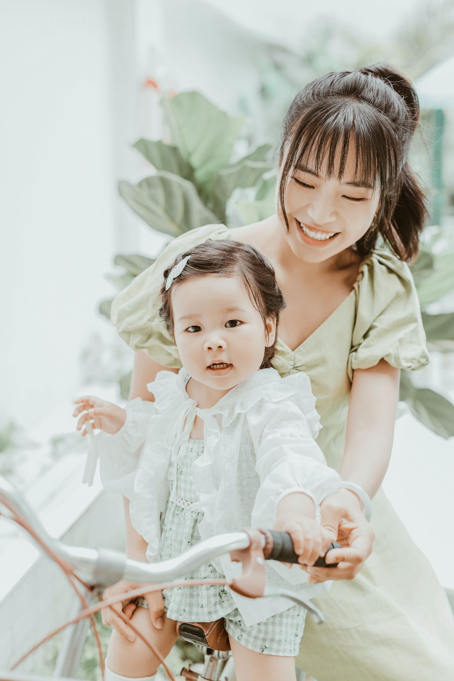 Asian mother in green dress smiles while helping her young daughter ride a bicycle outdoors.