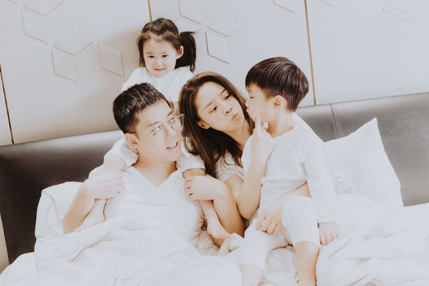 A loving Asian family of four laughs and cuddles together on a white bed in a bright room.