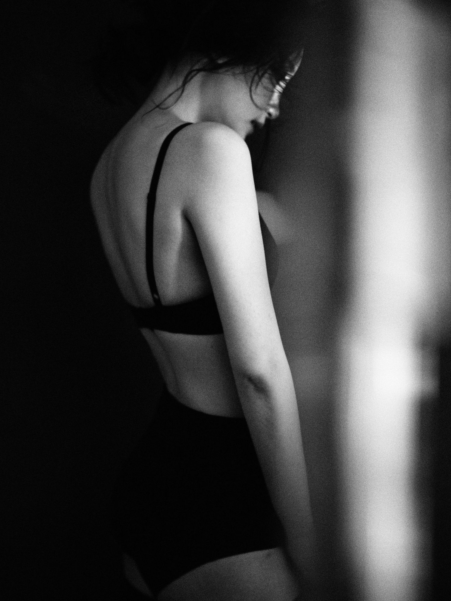 Black and white artistic photo of a woman in a black bralette and underwear viewed from behind in dim lighting.