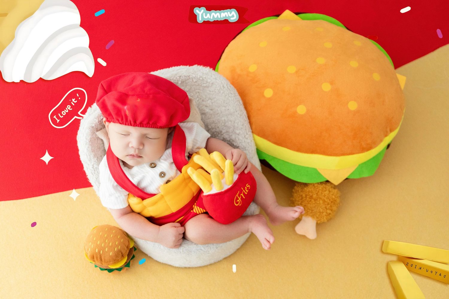 Adorable baby dressed as a chef holding toy fries, surrounded by burger props on a colorful red and yellow backdrop.