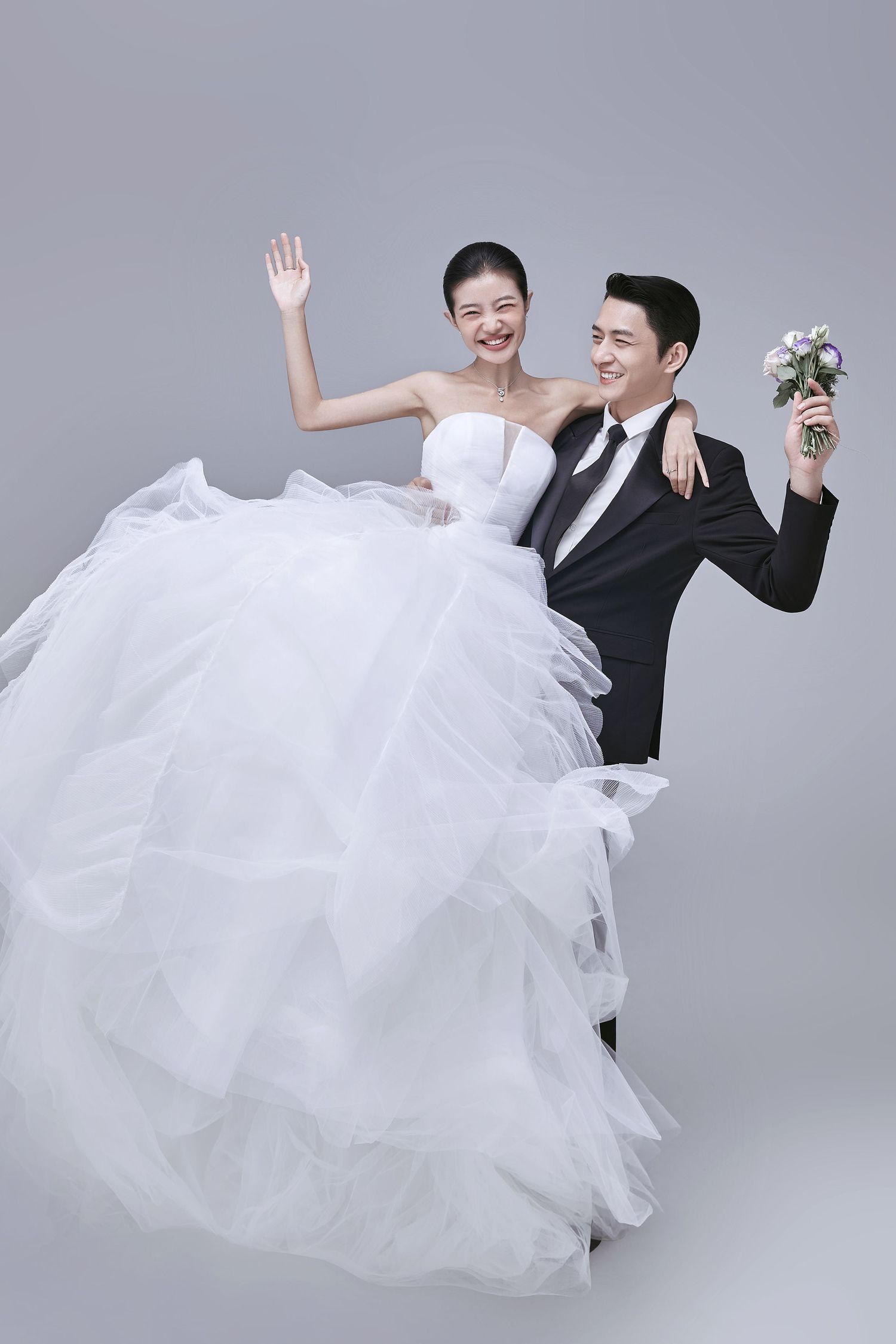 A happy bride and groom in wedding attire wave and celebrate together against a gray background.