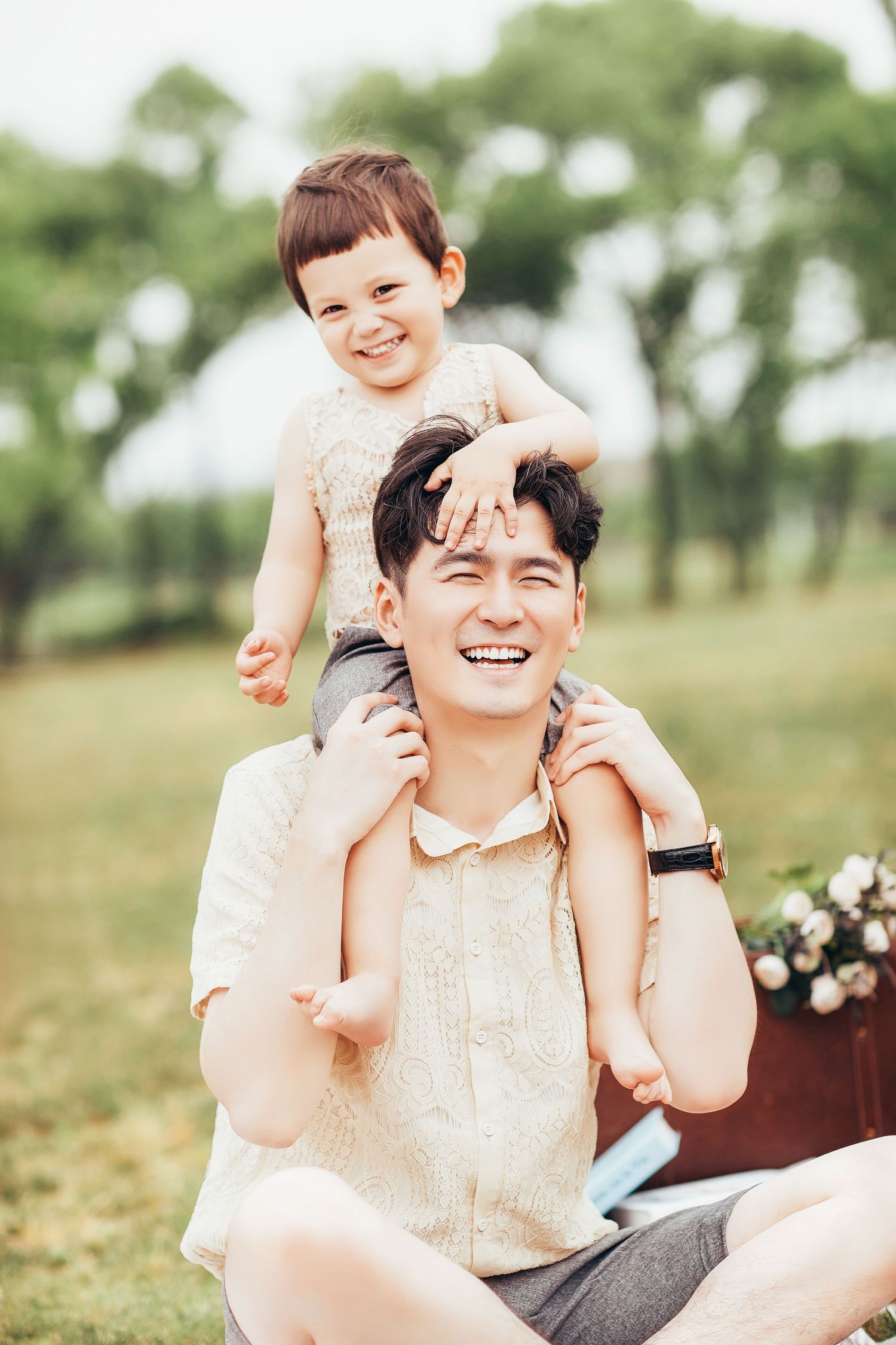 A laughing Asian father sits on grass with his young toddler perched happily on his shoulders.