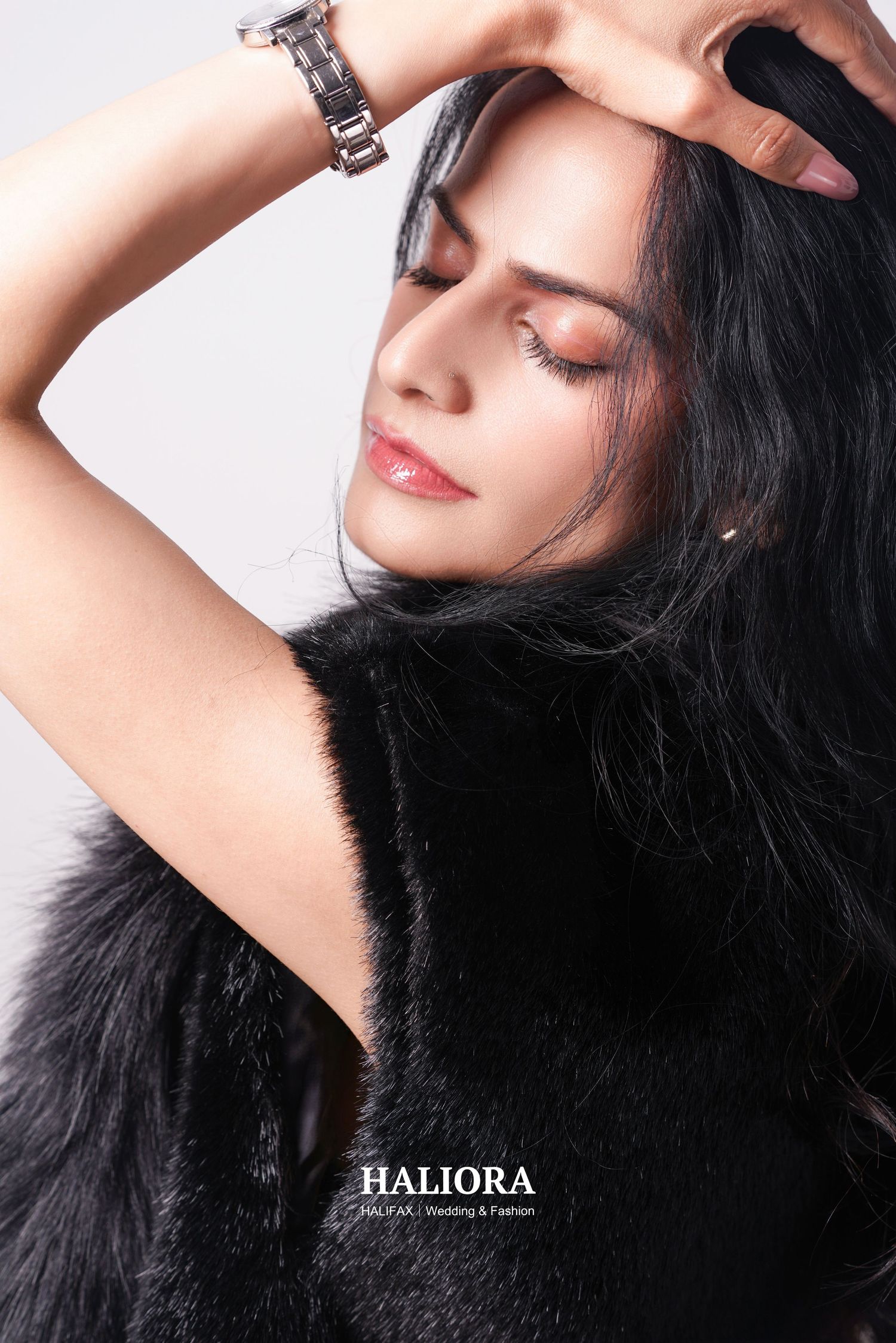 Woman in black fur vest with eyes closed, hand raised to head, wearing silver watch, fashion portrait.