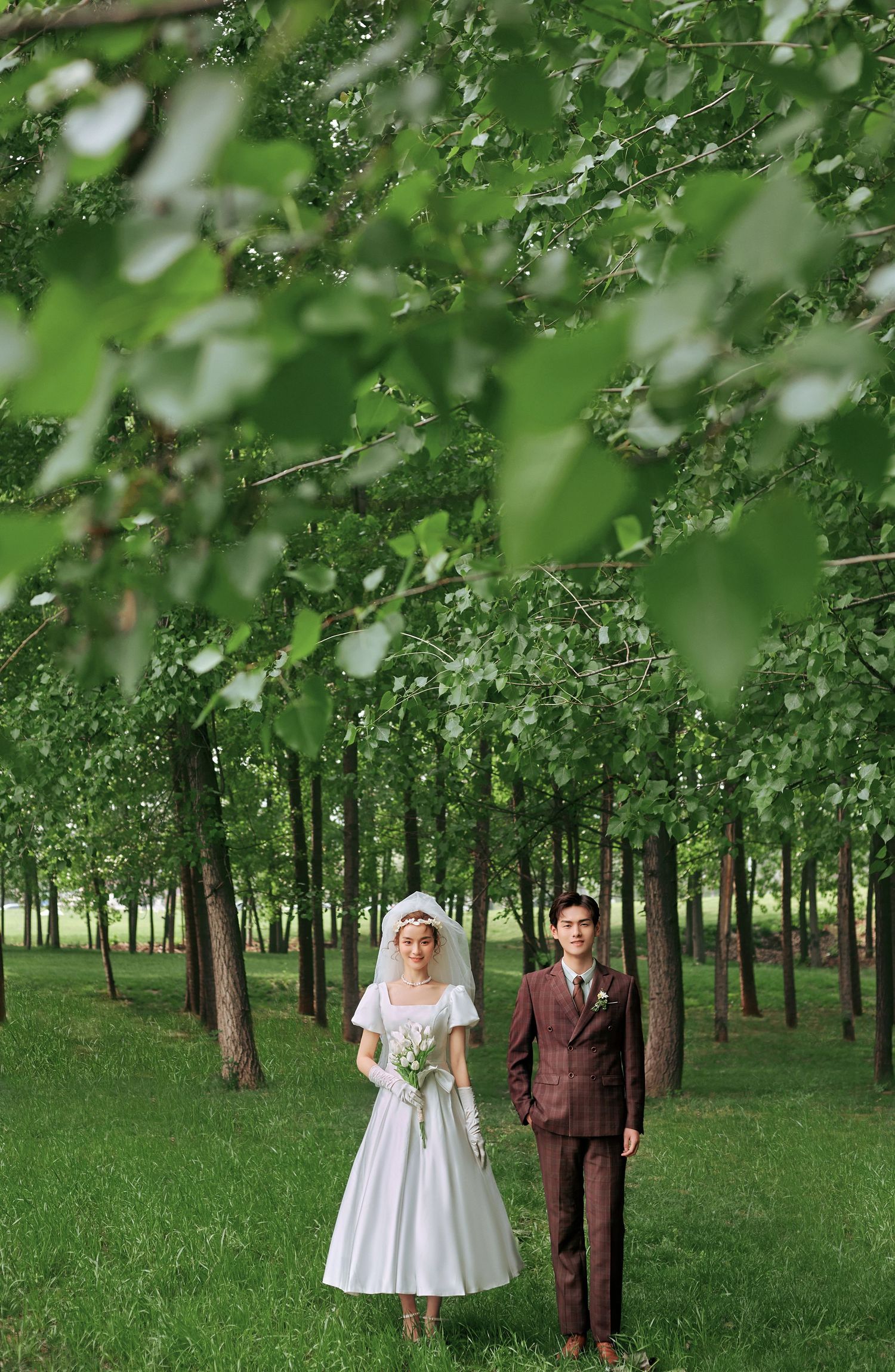 Bride and groom standing among tall green trees in lush forest during outdoor wedding photography.