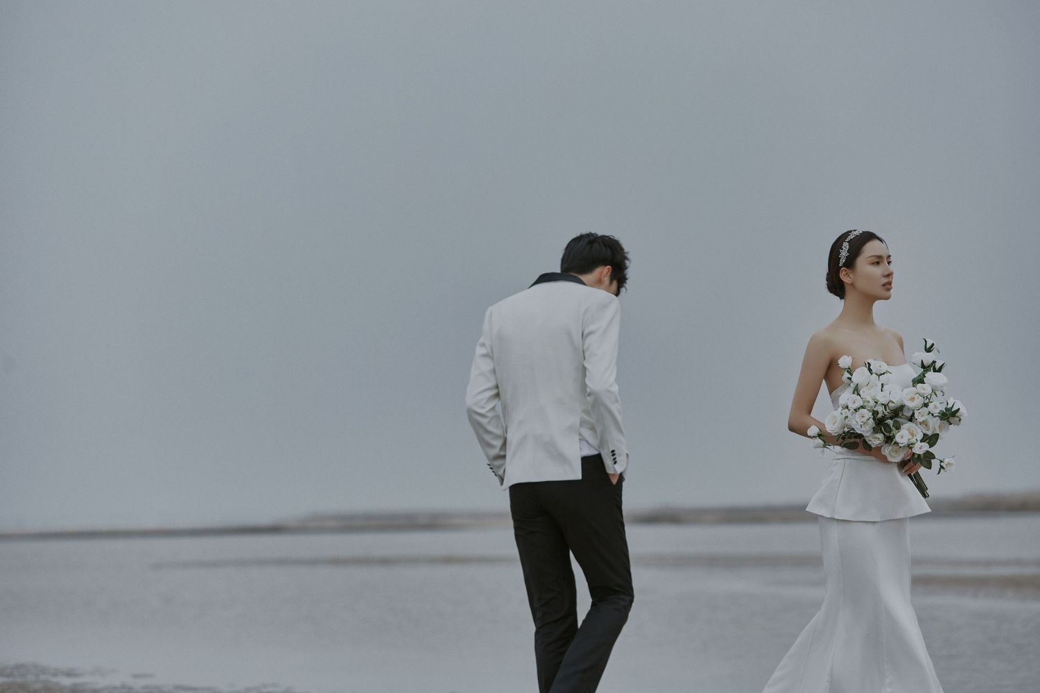 Bride in sleek white gown holds bouquet while groom stands nearby on a calm misty beach.