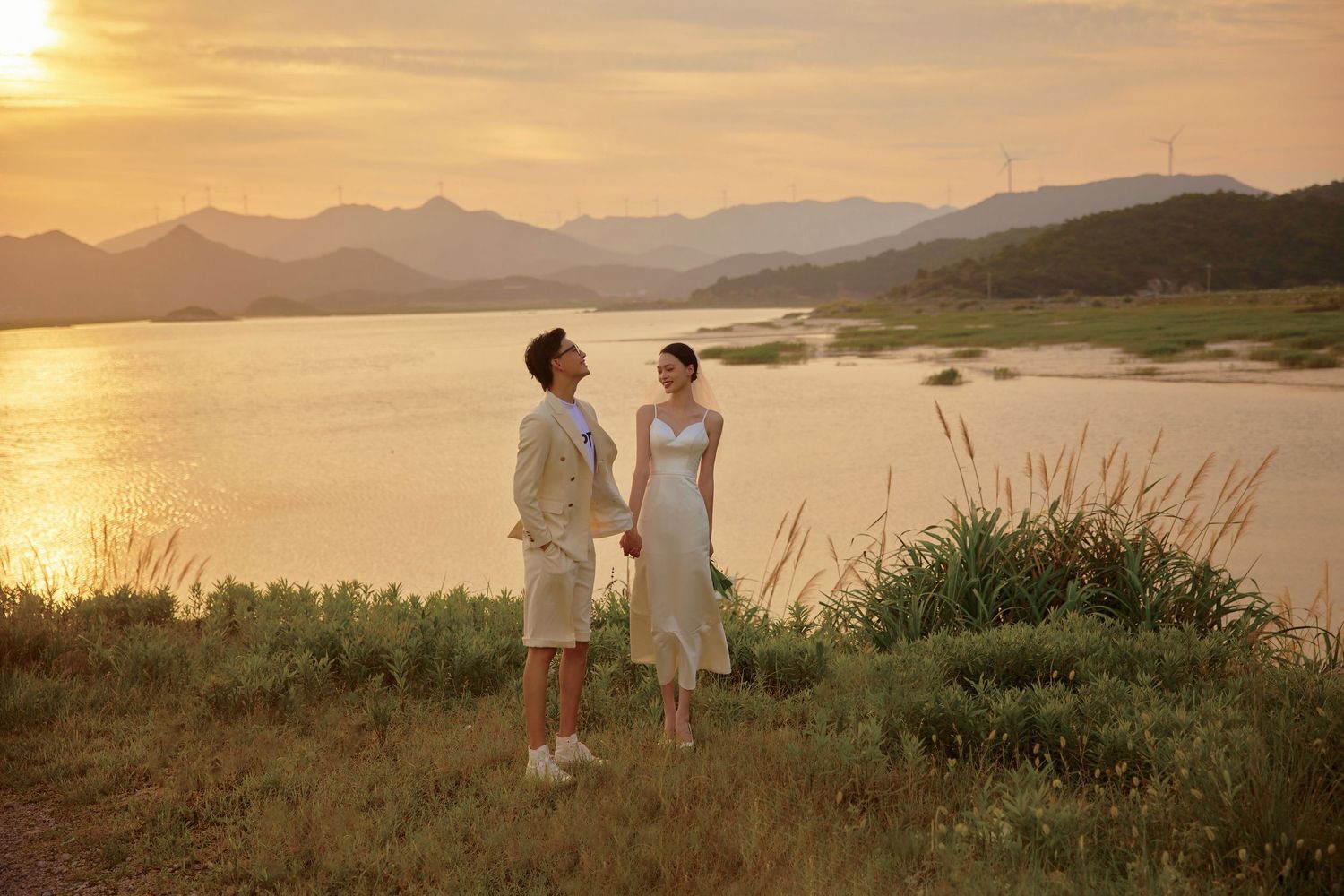 Couple holding hands by a scenic lake at golden sunset with mountain backdrop.