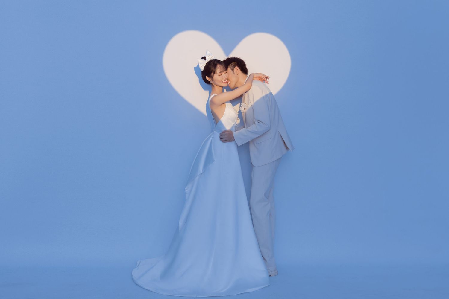 Couple in wedding attire embracing in front of a glowing heart backdrop on a blue background.