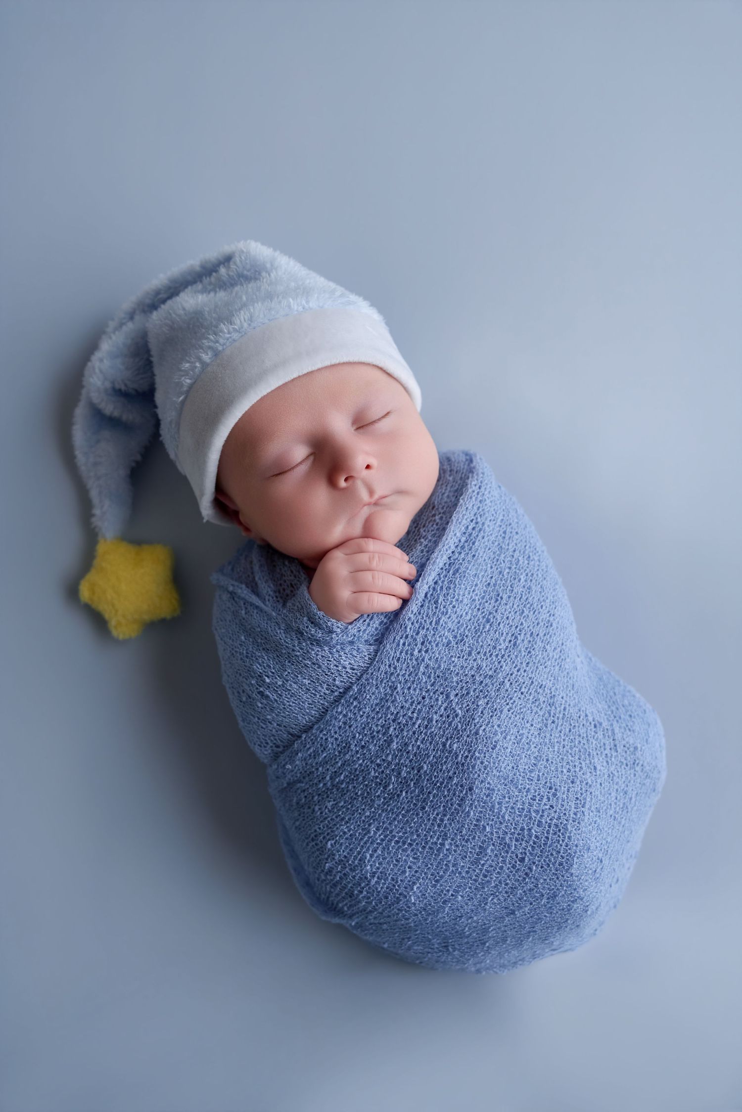 Sleeping newborn baby swaddled in blue knit wrap wearing a nightcap with yellow star, lying on blue background.