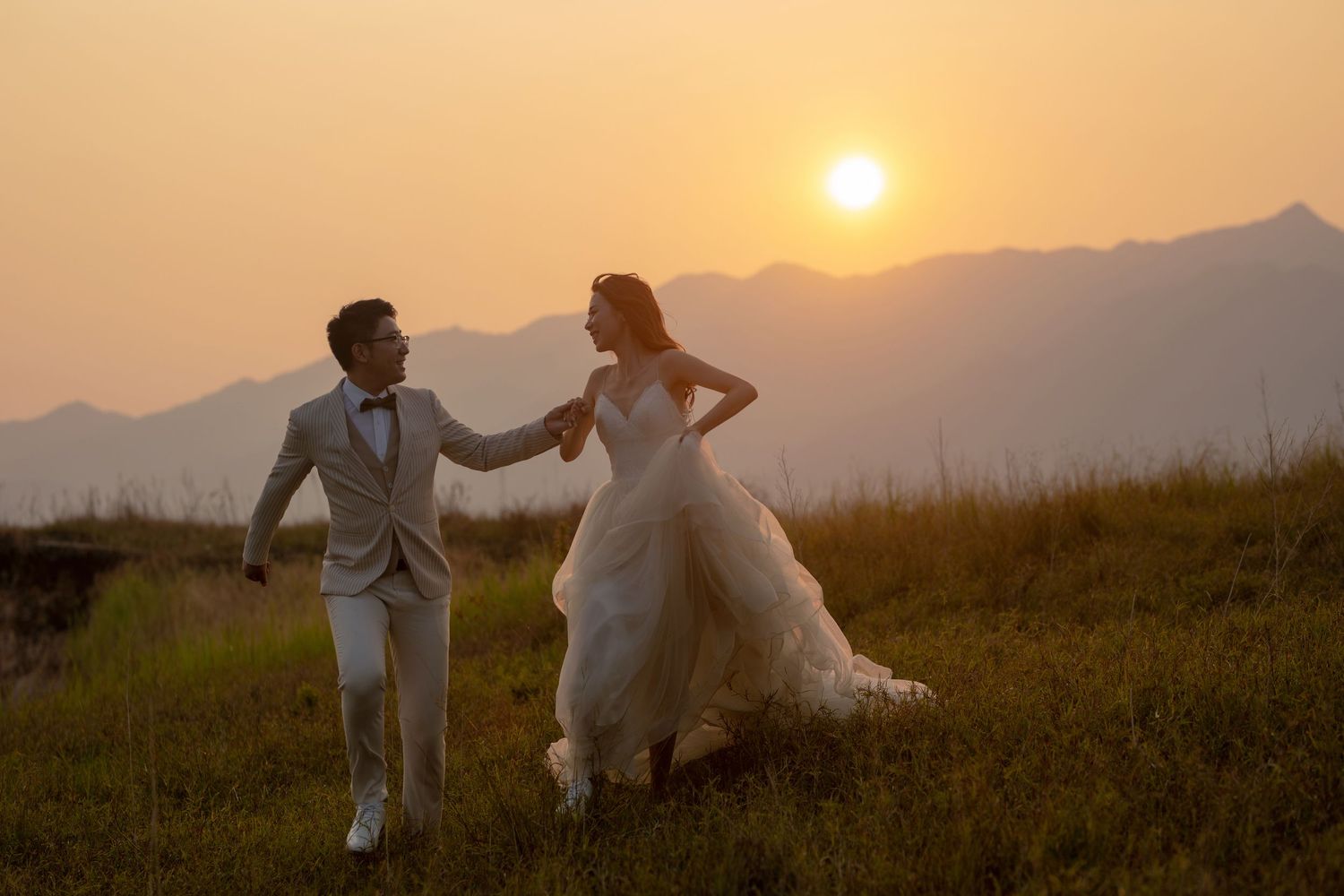 Romantic wedding couple walks hand in hand through a grassy meadow with breathtaking golden sunset and mountains behind.