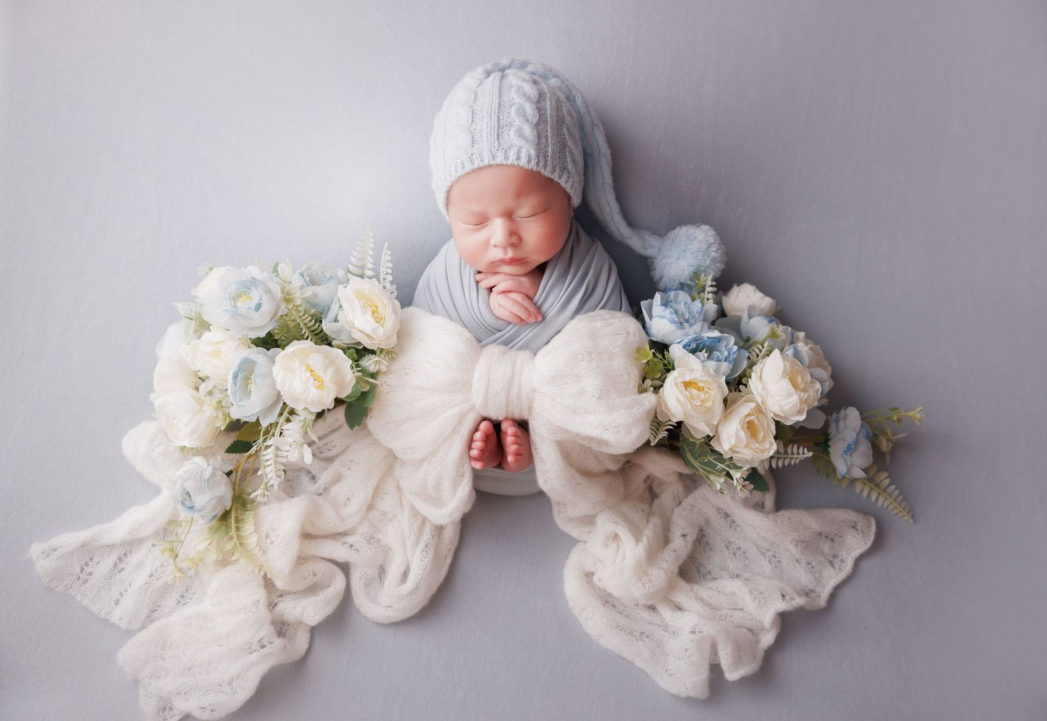 Swaddled newborn baby in blue knit hat sleeping with white bow and floral bouquets on soft gray background.