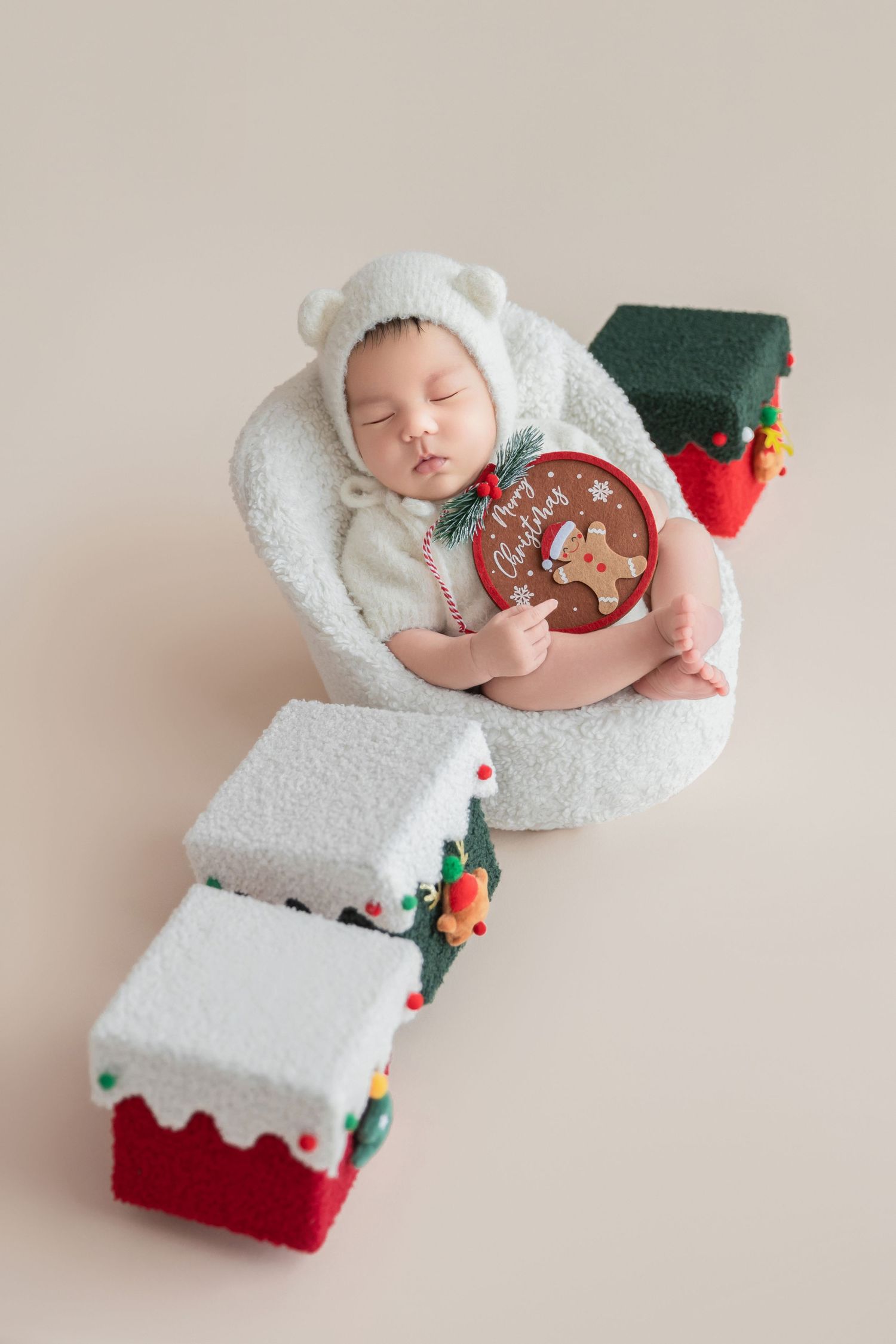 Sleeping newborn baby in white bear outfit holding Christmas ornament beside decorative miniature holiday houses on beige background.