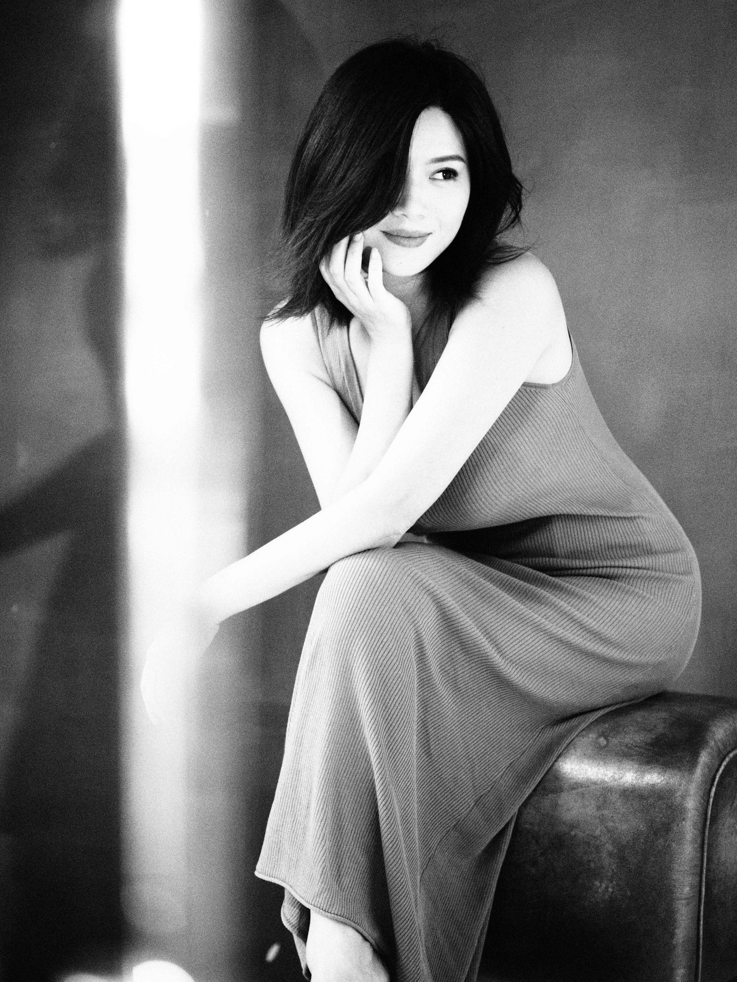 Black and white photo of an Asian woman in a slip dress seated gracefully on a leather chair in soft light.