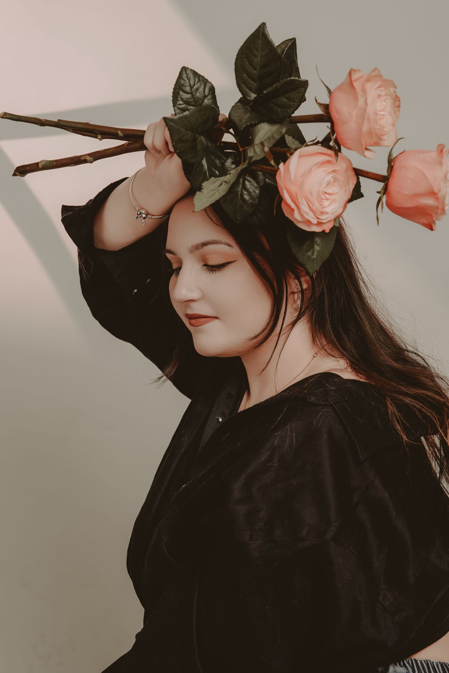 Young woman in black outfit holds pink roses above her head with eyes closed against a neutral background.
