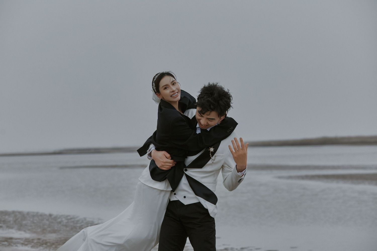 Joyful couple in black and white wedding attire playfully pose on misty beach shoreline.