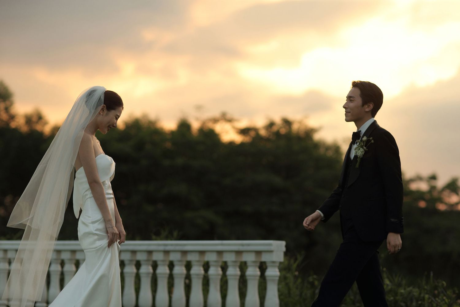 Bride in flowing white gown and veiled groom walk toward each other on terrace at sunset.