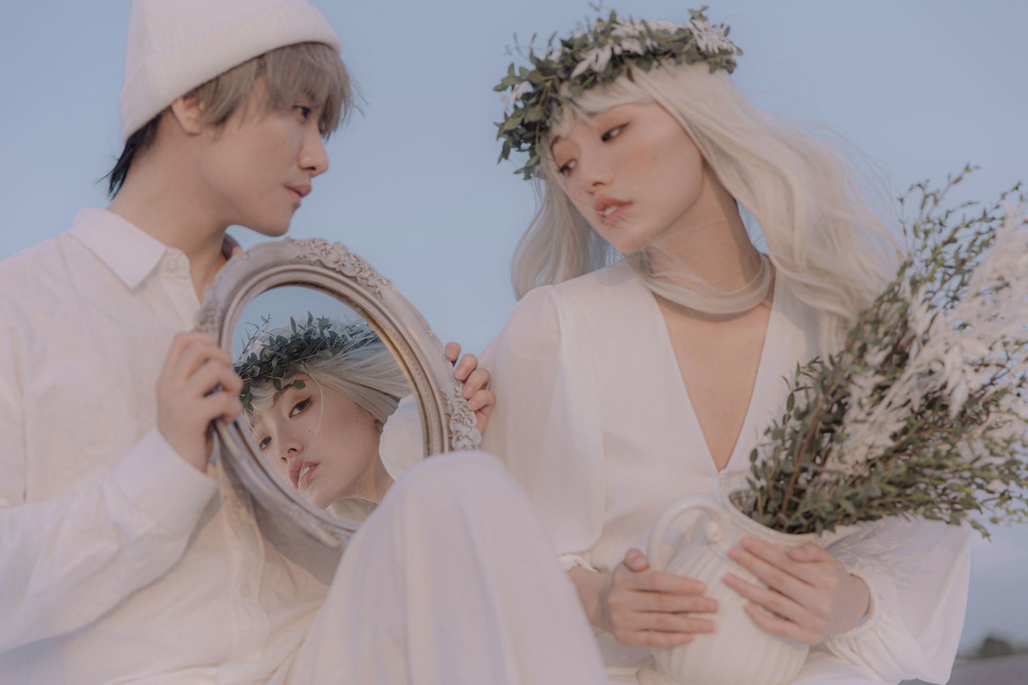 Two people in white outfits pose with a round mirror outdoors, woman wearing floral crown holding dried flowers.