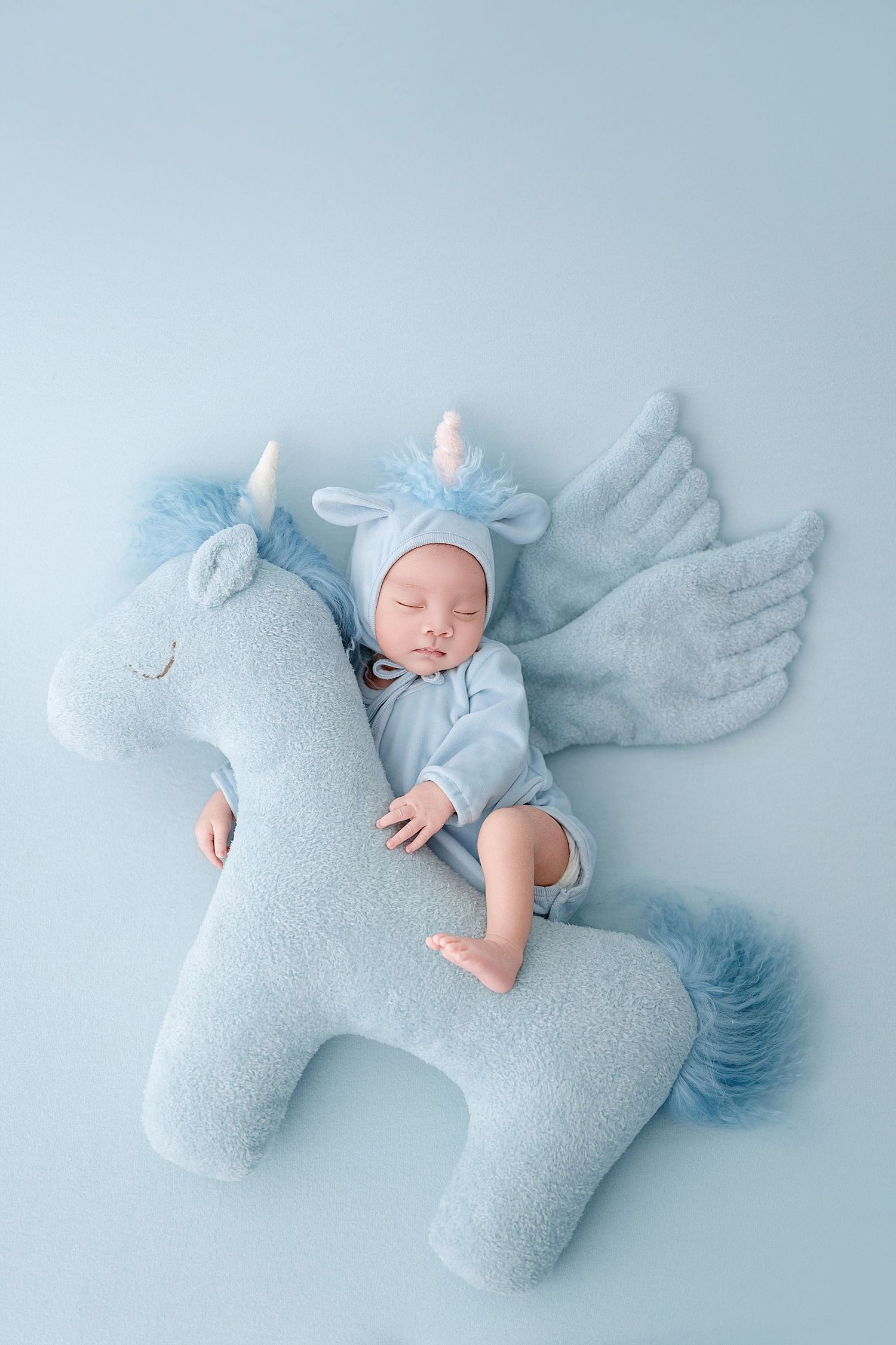 Sleeping newborn baby in blue unicorn costume lying on matching plush unicorn toy against light blue background.