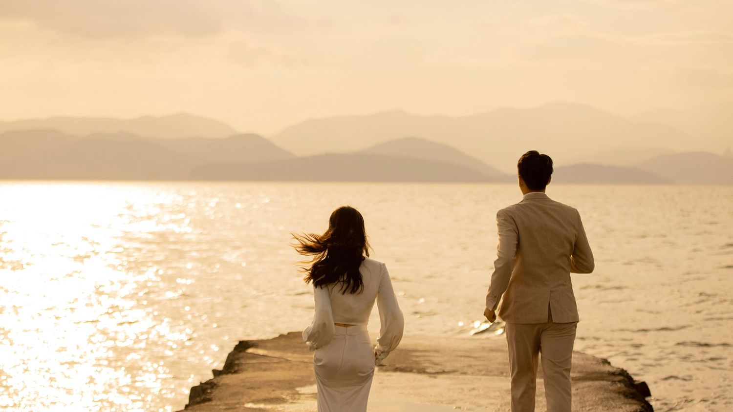 Wedding couple walking on seaside pier at golden sunset with mountain views in background.